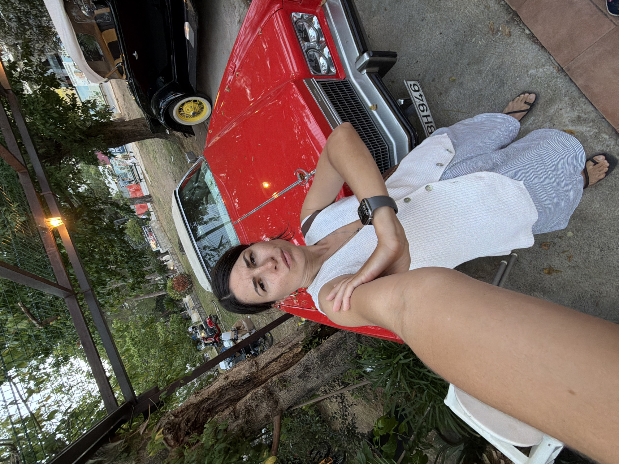 This rotated selfie captures a woman posing in an outdoor setting, likely a parking area or yard in Budva, Montenegro. The central subject is the woman, who looks directly at the camera with a neutral expression. She has dark hair pulled back and is dressed in a white, button-down sleeveless top, blue and white striped wide-leg pants, and black flip-flops. A dark smartwatch is visible on her left wrist.
Prominently featured alongside her is a bright red classic American car, possibly from the 1970s or 80s, showcasing its chrome bumper, large grille, and dual headlights. Its front license plate displays the text "BH 976HB". Partially visible behind the red vehicle is another classic car, black in color, notable for its distinctive yellow wire wheels and whitewall tires.
The background consists of lush green trees, including a large fallen log or tree trunk. A metal fence structure and hints of distant buildings suggest an urban or semi-urban environment. The ground is paved concrete. The lighting suggests it is daytime, possibly late afternoon, with soft, natural light illuminating the scene. The activity is a casual selfie, capturing a relaxed moment with vintage automobiles.