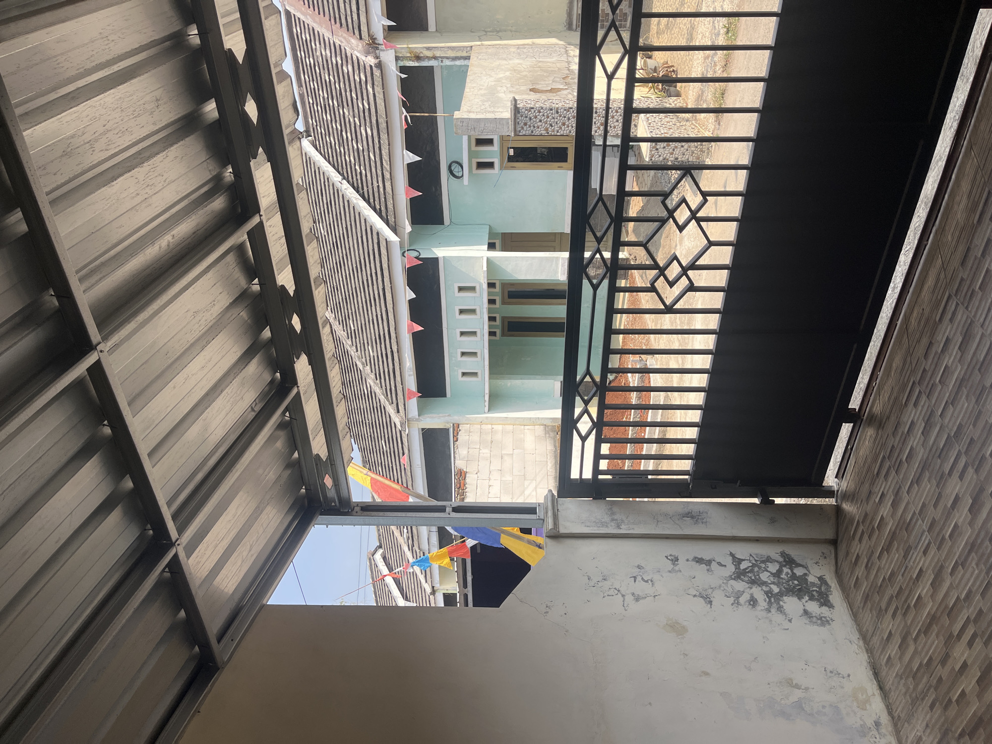 The image depicts a street view from a covered porch in Cirahayu, Indonesia. A corrugated metal roof is visible overhead, supported by metal beams. To the left, a weathered, off-white wall with peeling paint and water stains occupies the foreground. The floor is tiled with a pattern of brown and gray rectangles. Beyond the porch, a black metal gate with diamond-shaped accents separates the property from the street.
Through the gate, several houses are visible. The nearest house on the right has a light green facade and a textured stone pillar. A string of red and white triangular flags decorates the roofline of this house and extends to the neighboring house on the left, which is light blue and features a series of rectangular window openings. Two tall, colorful banners, one yellow and red, the other blue and yellow, are partially visible, hanging from above. The background shows more houses with gray tiled roofs and a clear blue sky. The street appears to be unpaved. The scene suggests a residential area, possibly decorated for a local celebration or event. No people are visible in the image.