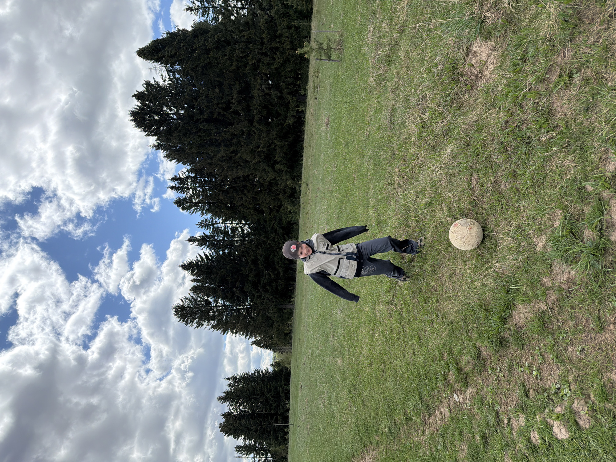 A medium-angle shot shows a person standing on a grassy hill next to a soccer ball. The person is wearing a brown hat, a tan vest, a black long-sleeved shirt, and dark pants. The grass is green and patchy. In the background, there are tall, dark green pine trees. The sky is blue with puffy white clouds. The location cannot be determined from the image.