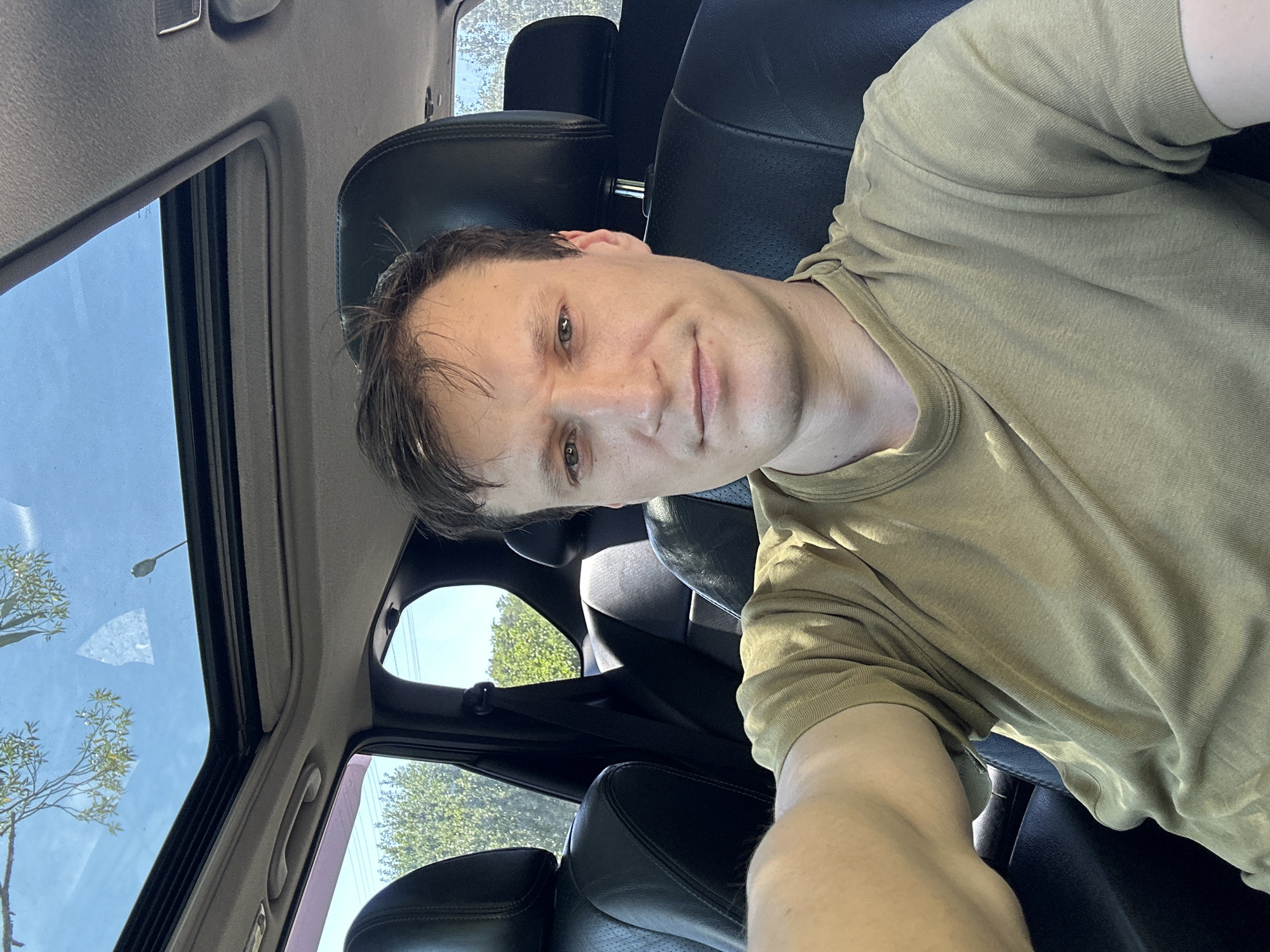 A man is sitting in the driver's seat of a car with a sunroof open, on a sunny day. He is wearing a green t-shirt and looking directly at the camera. The car's interior is visible, including black leather seats and the dashboard. Outside the car, trees and a clear blue sky are visible through the open sunroof and windows. The location is specified as Makiv, Ukraine.