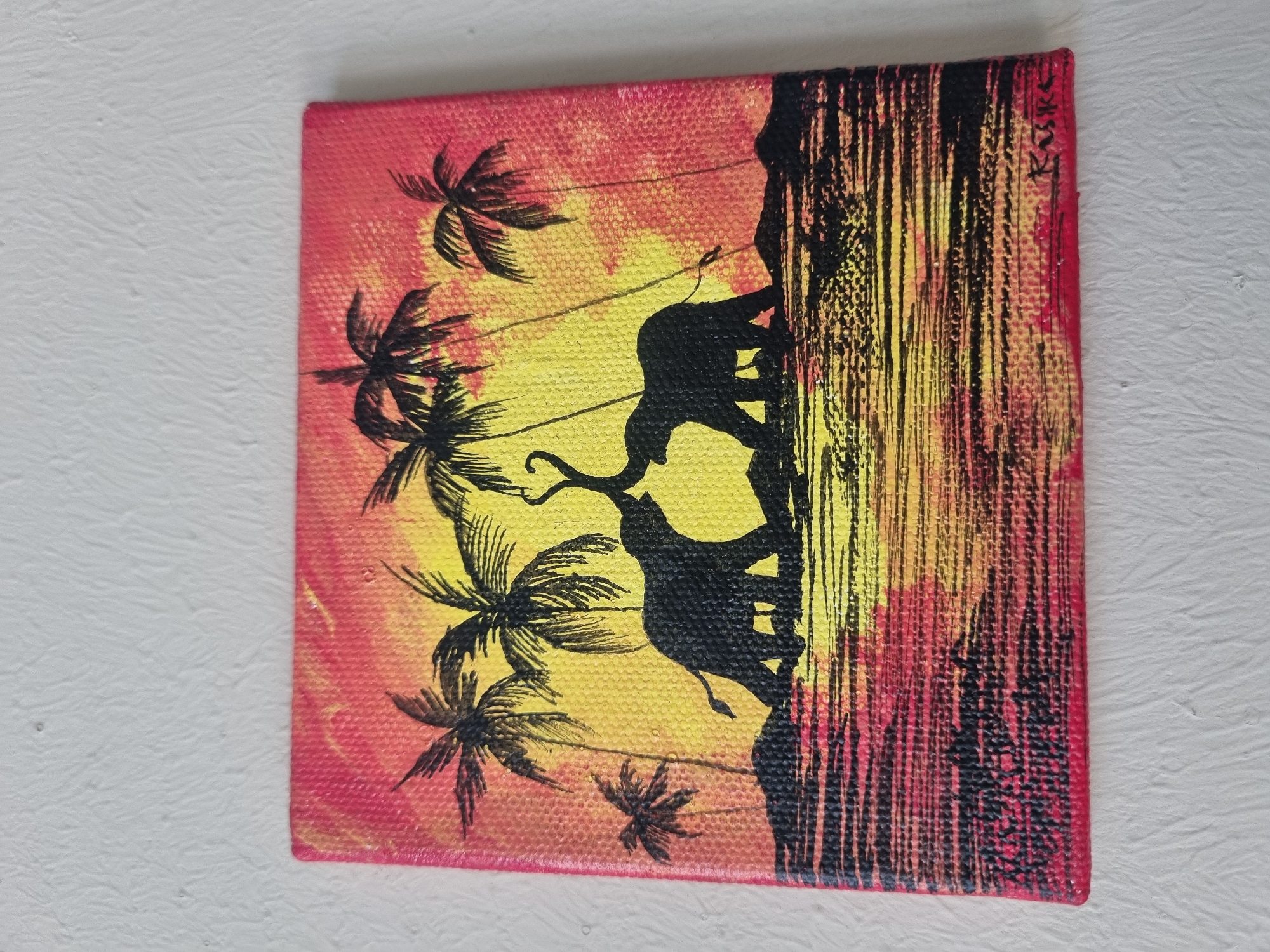 The image is a small, square painting on canvas, depicting a scene with silhouettes of elephants and palm trees against a gradient background of red and yellow, resembling a sunset or sunrise. On the right side of the painting, a vertical, textured black and yellow pattern suggests either a cliff face or dense foliage. The artwork features a signature "KuKu?" on the right edge. The subject matter and style do not definitively place the scene in Sochi, Russia, though tropical or semi-tropical elements are present.