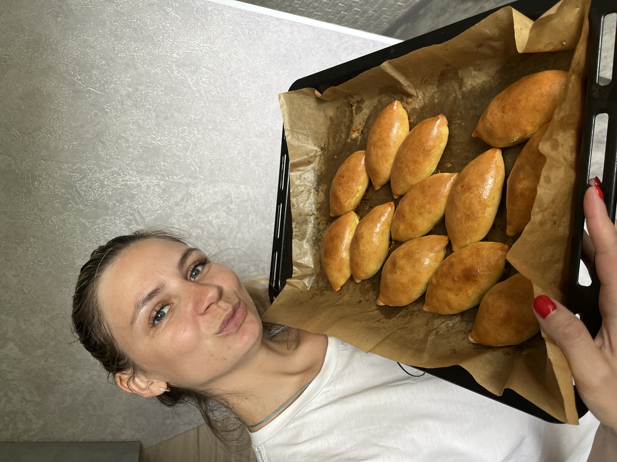 A young Caucasian woman with blonde hair tied back in a braid and blue eyes is holding a baking sheet filled with golden-brown, oval-shaped pastries. She is wearing a white t-shirt and a light blue necklace. Her fingernails are painted red. The pastries are arranged on brown parchment paper on a black baking sheet. The background consists of a textured, light gray wall. The scene appears to be indoors, possibly in a kitchen. There is no visible indication of the specific location within the city of Chaikovsky, Russia.