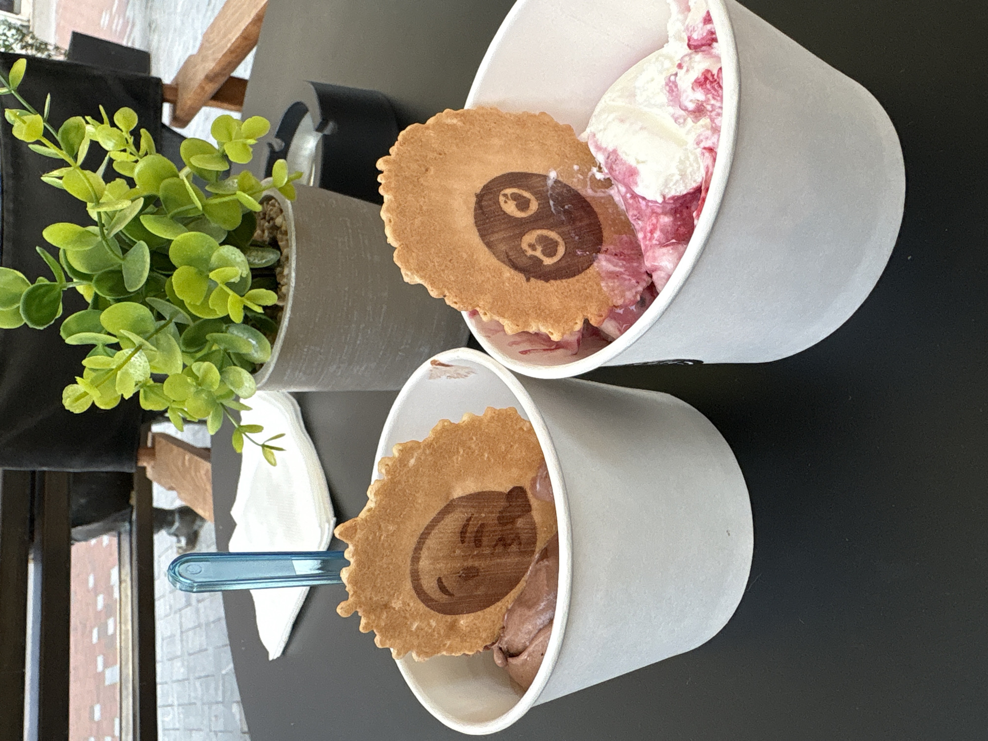 This image shows a close-up, angled view of two paper cups filled with ice cream, sitting on a black table outdoors. Each cup is topped with a thin, round wafer cookie imprinted with a cartoon face. The cup on the top right contains a mix of white and pink ice cream, while the cup on the bottom left contains brown ice cream. Beside the bottom cup is a blue spoon and a folded white napkin. A potted plant with green leaves sits to the left of the cups. The background shows a glimpse of a building facade, hinting at an urban setting, possibly Varna, Bulgaria. The composition suggests a casual, outdoor setting like a cafe or ice cream parlor.