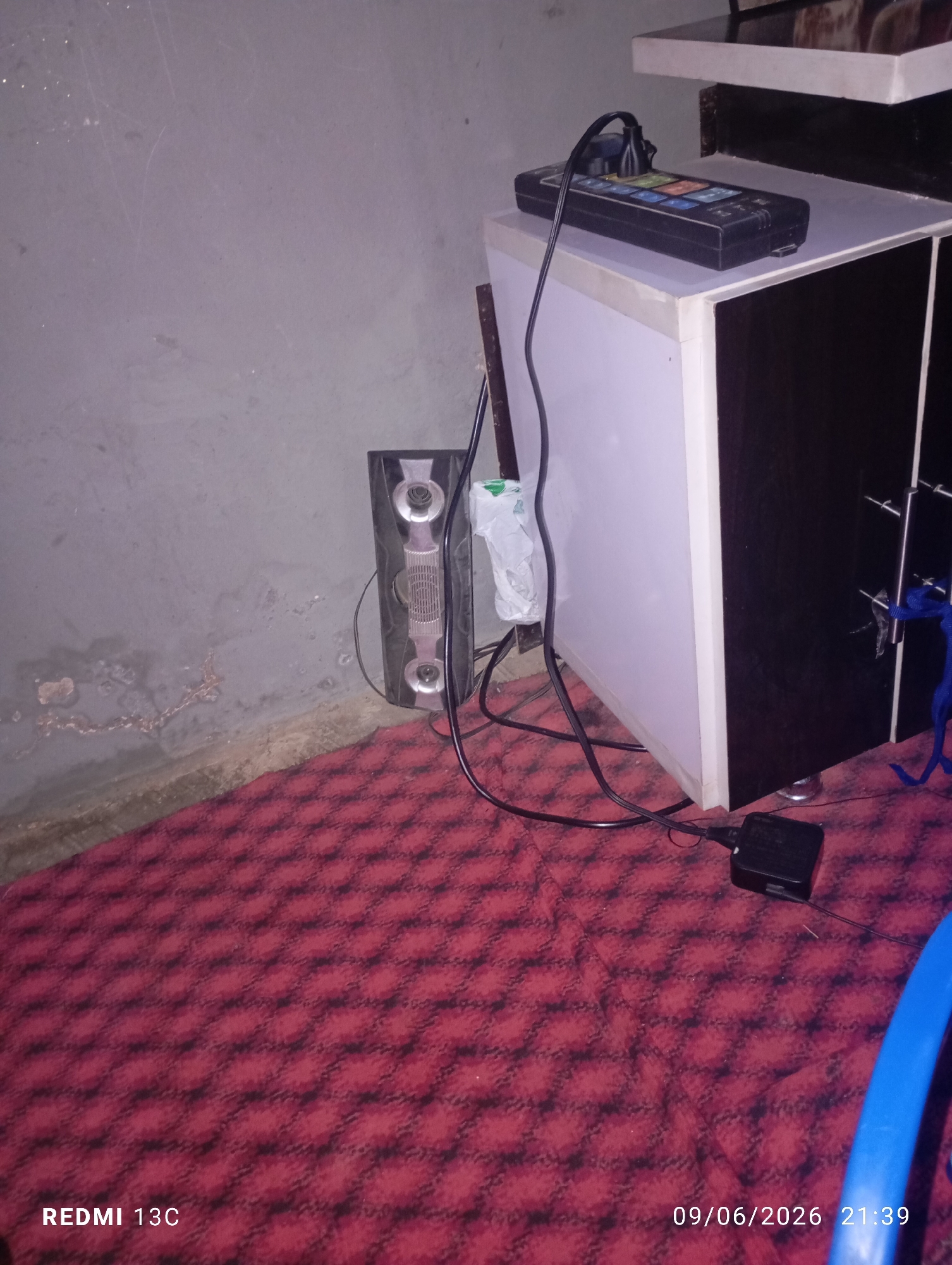 In an indoor scene with a gray, textured wall on the left and a red patterned carpet on the floor, a tall, black speaker with silver accents sits against the wall. To the right of the speaker, a white and brown cabinet is partially visible. On top of the cabinet, a black power strip with multiple outlets is plugged in, with a black cord extending from it. Another black cord snakes across the floor, connecting to a small black adapter. A white plastic bag is attached to the side of the cabinet. A blue cylindrical object is visible on the far right of the frame. The timestamp "09/06/2026 21:39" and the text "REDMI 13C" are present in the lower portion of the image.