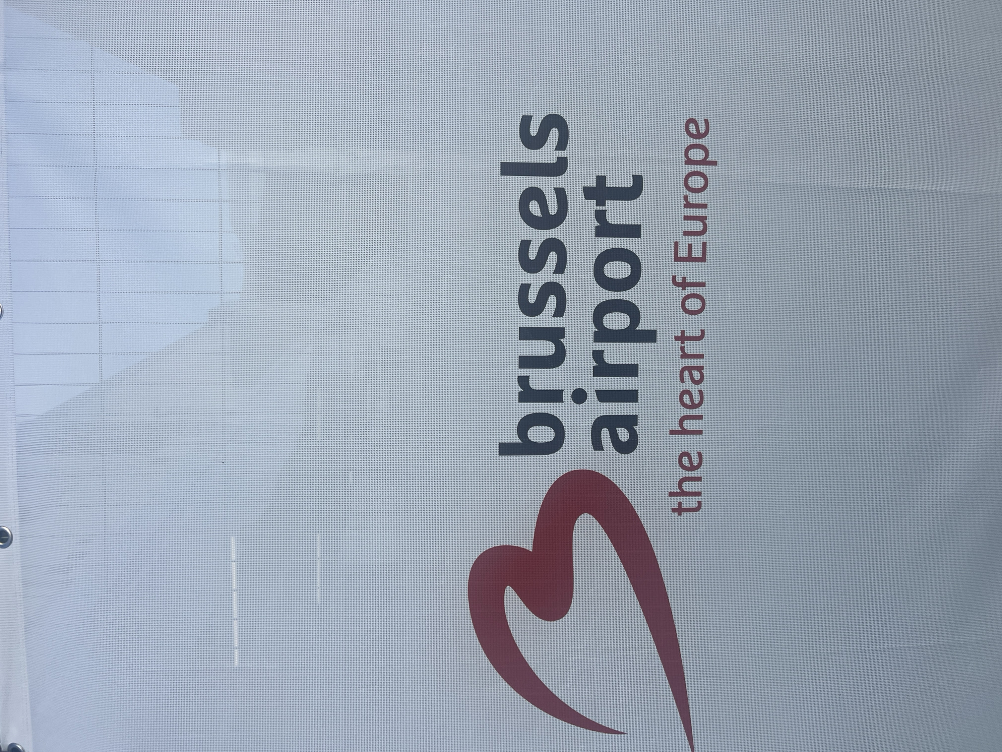 A white banner with the Brussels Airport logo and slogan is displayed. The logo features a stylized red "3" on the left side, to the right of which is the text "brussels airport" in dark gray. Below this, the slogan "the heart of Europe" is written in red. The banner is suspended outdoors, as indicated by the bright sky and building reflections visible through the mesh fabric. The location is identified as Brussels Airport in Zaventem, Belgium.