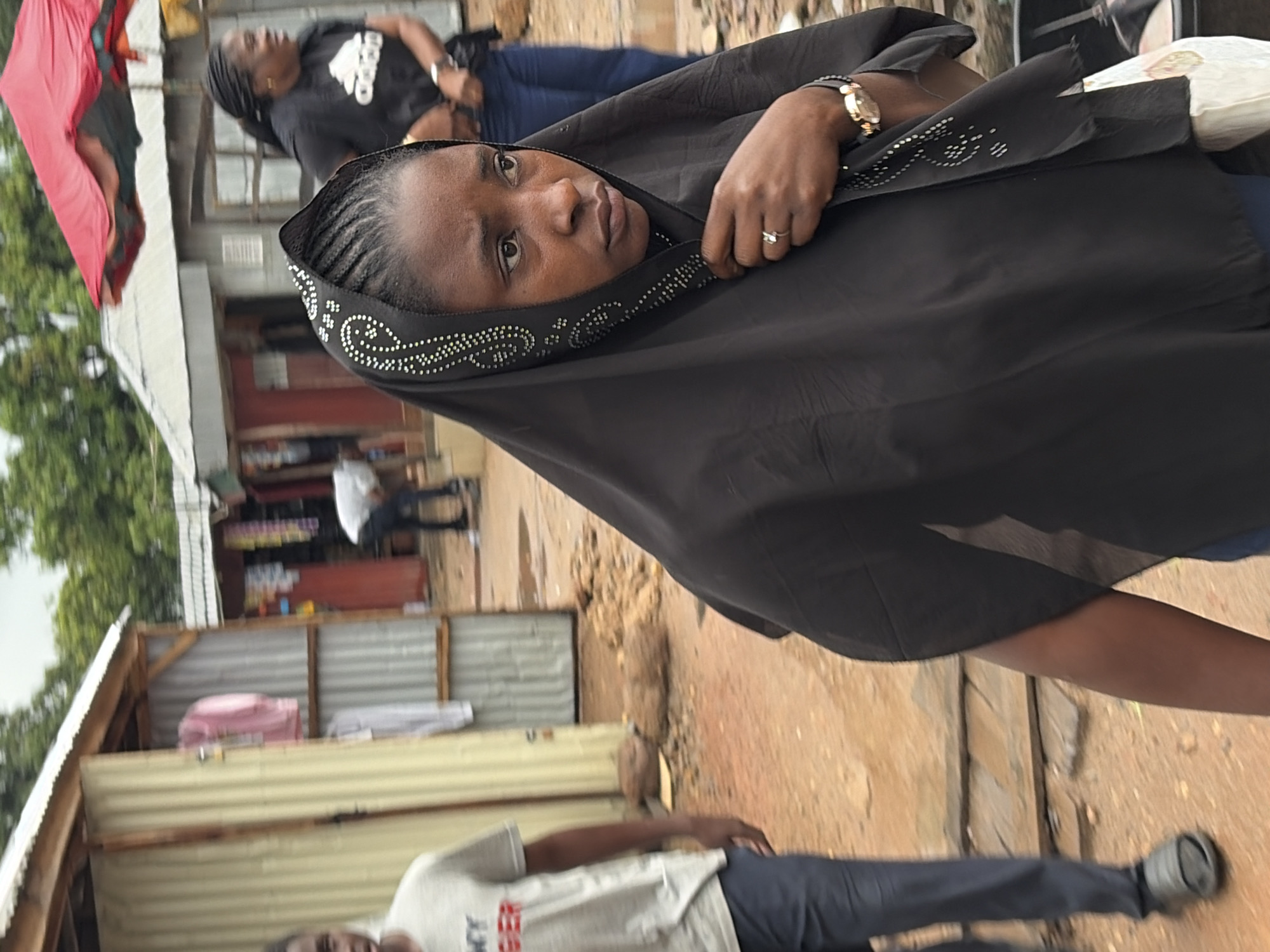 The image depicts an outdoor market scene, likely in the city of Jos, Nigeria. In the foreground, a woman wearing a black hijab with decorative white embellishments is partially visible. She is looking to her right with a concerned expression. A ring is visible on her left hand, and she is wearing a wristwatch. To her left, a young man wearing a grey t-shirt with red lettering and dark pants is walking away from the camera. Behind them, several stalls with corrugated metal roofing and walls are visible. One stall displays pink and white clothing items. Further in the background, another woman in a black t-shirt with an Adidas logo is visible, as is a red market umbrella. The ground appears to be dirt and there are some scattered debris. The overall atmosphere suggests a busy, informal marketplace.