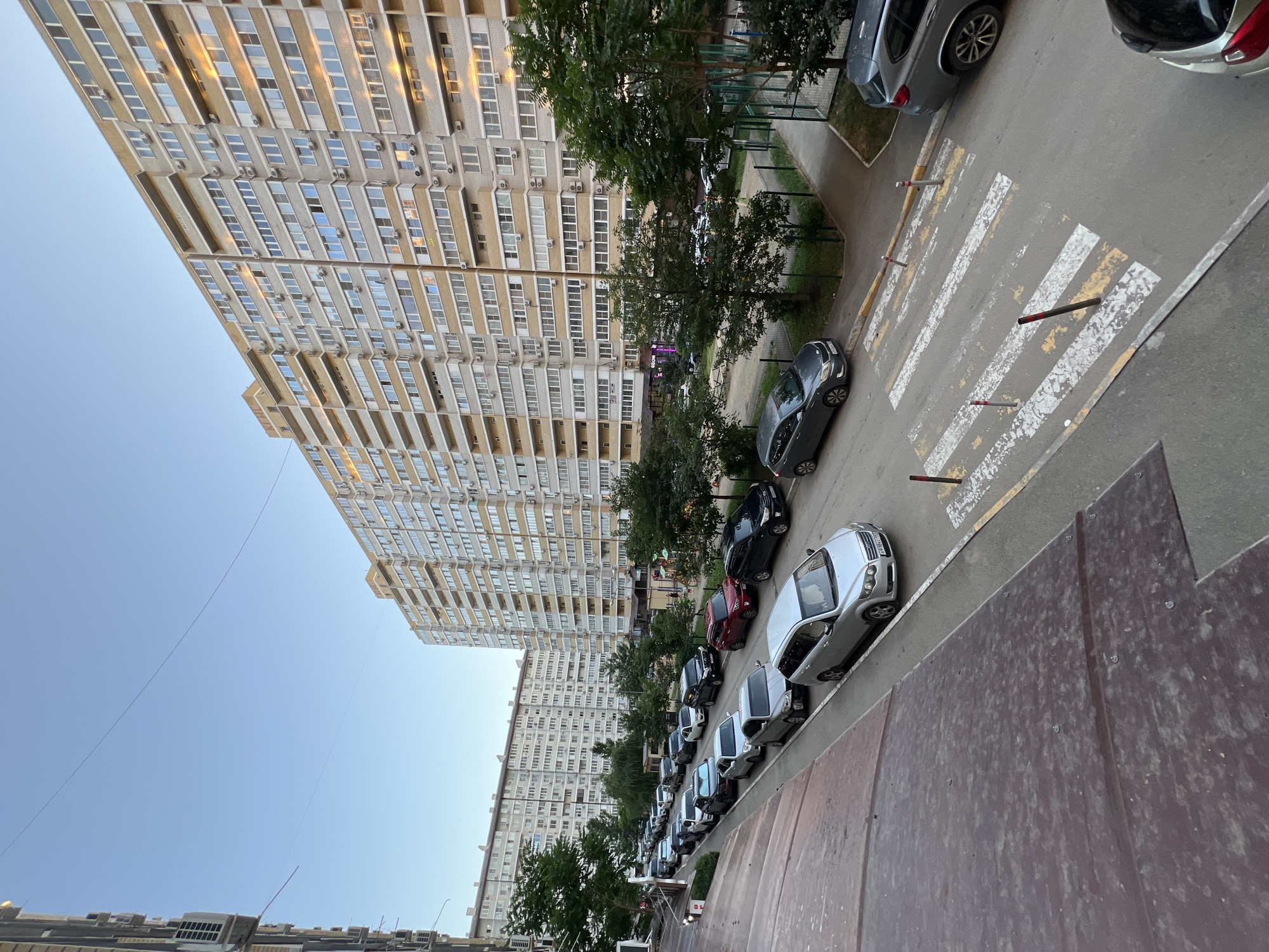 This media file depicts a street scene in Krasnodar, Russia. The primary subjects are a long, multi-story apartment building dominating the left side of the frame and a row of parked cars along the right side of a street. The apartment building is tall, with numerous windows and balconies, suggesting a residential area. The cars are parked in a line, some facing forward and others backward, indicating a typical urban parking situation.
The scene is outdoors, on a paved street with a marked pedestrian crossing. Trees are visible between the building and the street, providing some greenery. The sky is clear and blue, suggesting a daytime setting. The lighting suggests it might be late afternoon or early evening, as the sun casts warm reflections on the building's windows. There are no people visible in the frame, and no specific activity or event is taking place other than the general scene of urban life.
Notable details include the architecture of the apartment building, the variety of car models, and the condition of the pedestrian crossing, which shows some wear and tear. The overall impression is one of a common urban environment in Russia. No visible text is present in the image.