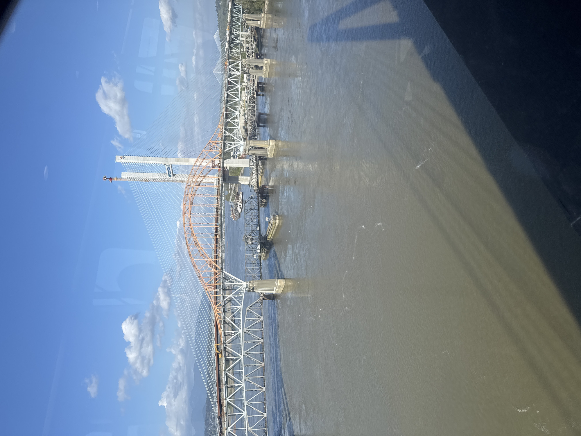 This image depicts a portion of the Pattullo Bridge and the under-construction New Pattullo Bridge spanning the Fraser River, as viewed from an elevated perspective, possibly from the sky or a high building in New Westminster, Canada. The old bridge is a through truss bridge with a gray-colored metal structure with concrete piers and is positioned diagonally. The new bridge, with its distinctive cable-stayed design, has two large, white towers with orange curved structural elements and many support cables. The sky is clear blue with some clouds. The river is murky and dark, with noticeable shadows. A partial view of a dark surface, likely from the window or structure from which the image was captured, is present in the right side of the image.