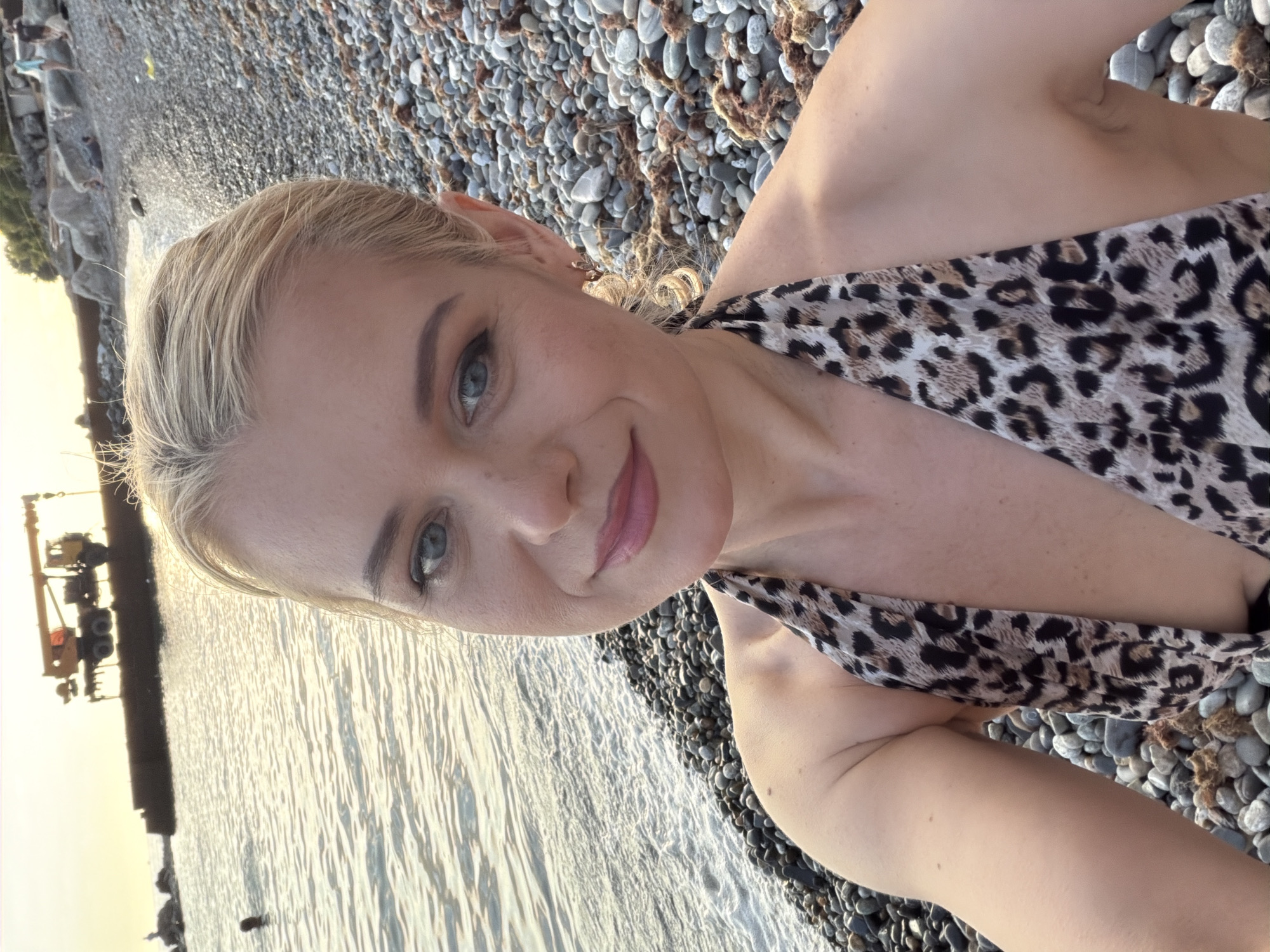 A blonde woman with blue eyes is taking a selfie on a pebble beach in Sochi, Russia. She is wearing a leopard print bikini. In the background, there is the sea and a large yellow crane on a pier. The sun is setting, casting a warm glow over the scene. There are other people in the background on the beach and in the water.
