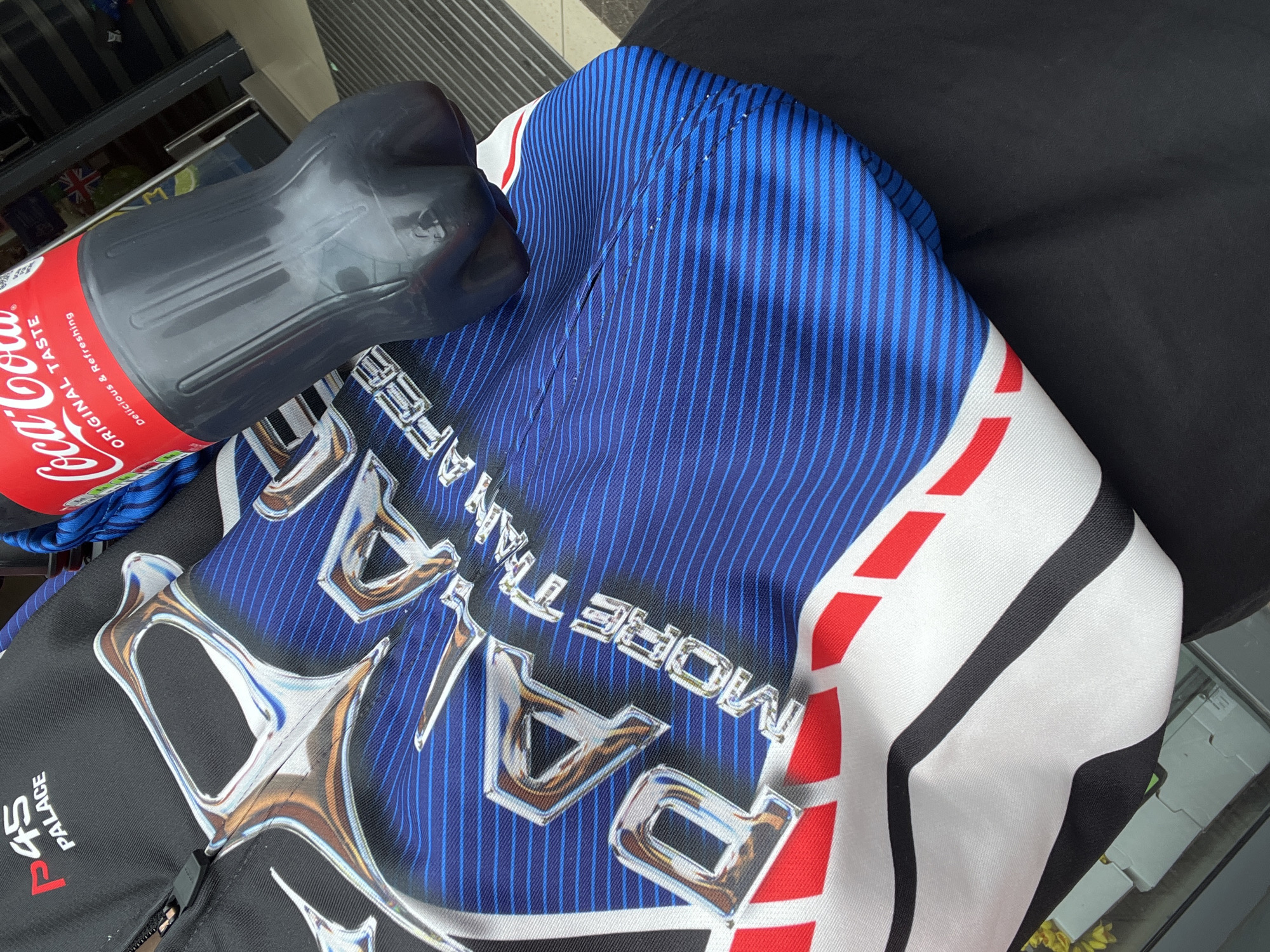 The image presents a close-up view of objects arranged on a black surface, possibly a lap or seat. A partially visible 1.75-liter bottle of Coca-Cola in a gray, ribbed plastic bottle with a red label rests on a folded or draped garment. The garment features a busy design, predominantly blue with thin vertical stripes, bold white stripes, and red accents. The word "PALACE" in large, reflective letters. A portion of the garment on the left has a black base with the word "PALACE" and the letter "P" stylized with the number "45" incorporated. The background indicates an indoor setting, likely a store or commercial space, evidenced by shelving units and glass surfaces. The location of this image cannot be confirmed based on what is visible.