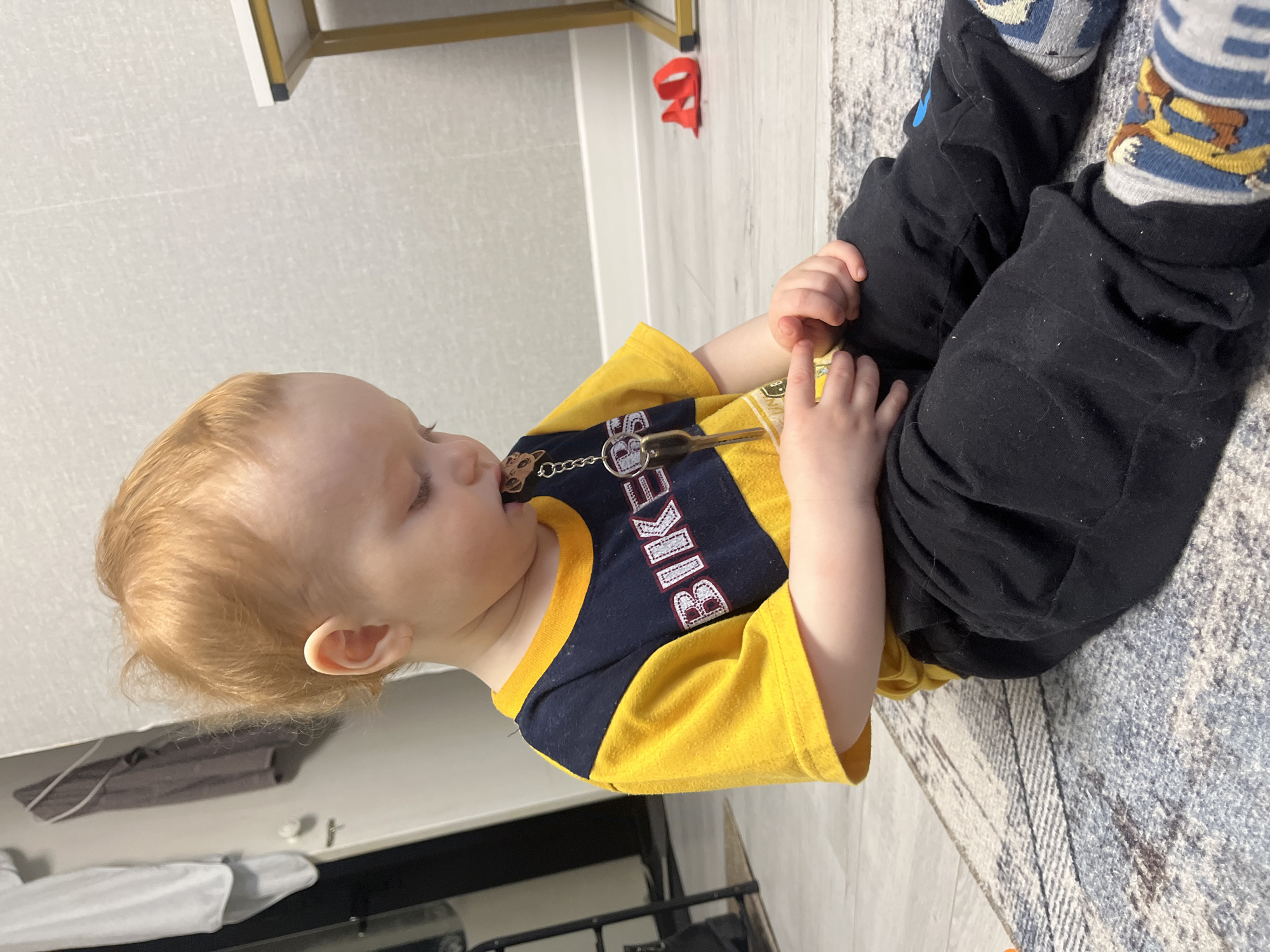 A toddler with red hair lies on a gray patterned rug in a room with light gray walls and flooring. The toddler wears a yellow and navy blue shirt with "BIKES" printed on it, black pants, and blue and gray socks with cartoon character patterns. The toddler holds a key ring with keys in their hands, near their mouth. A red object is partially visible on the floor near a white cabinet leg. A white table or countertop is visible in the background, along with what appears to be a black metal structure, possibly part of furniture. The location is not possible to determine based on the image.