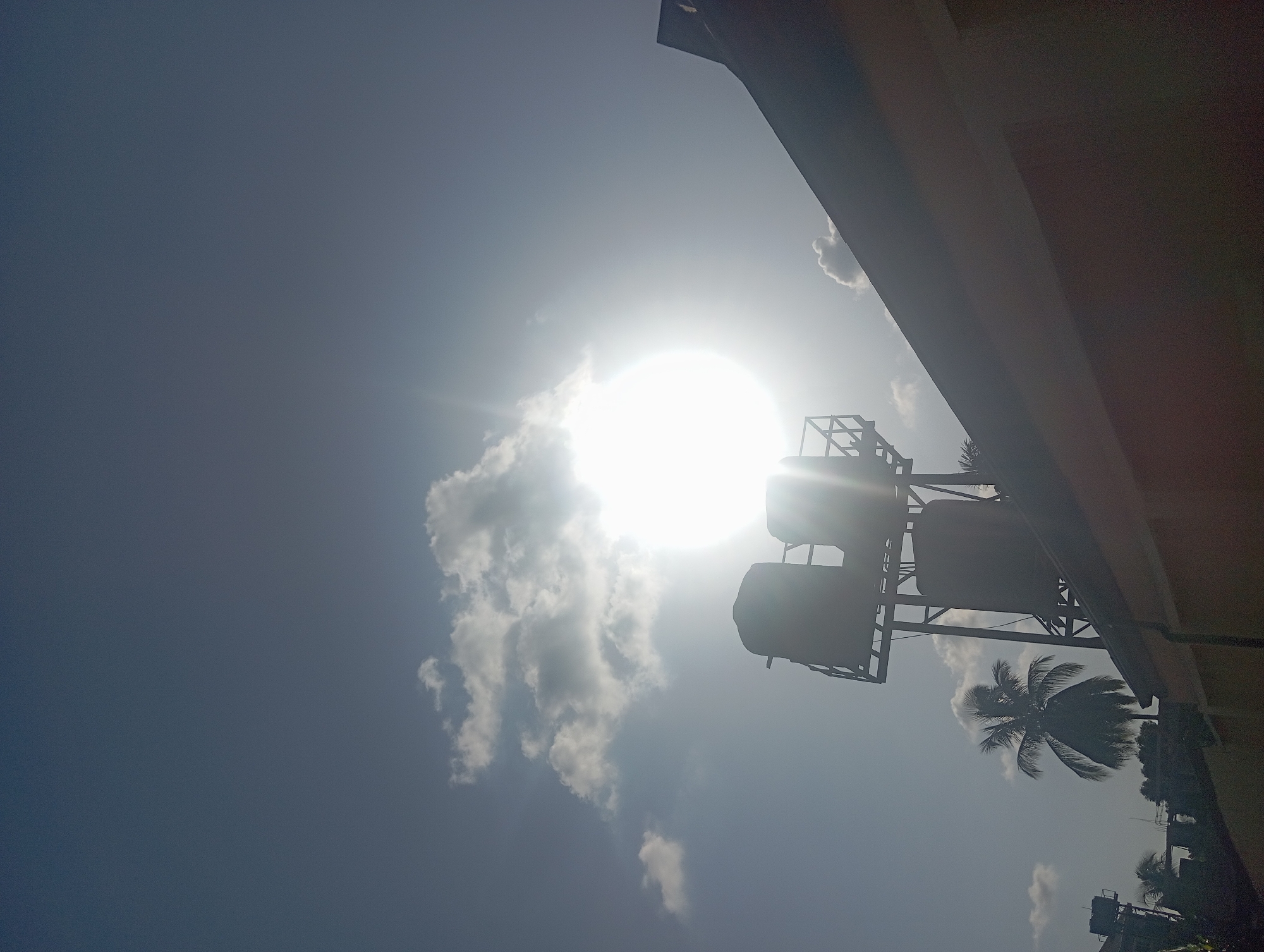 The image, apparently taken in Nkpor, Nigeria, shows a bright, sunny sky with scattered clouds. Silhouetted against the sky are: a support structure holding two water tanks, the edge of a building, and a palm tree. The angle suggests the photo was taken looking upwards from ground level. There are no people visible.