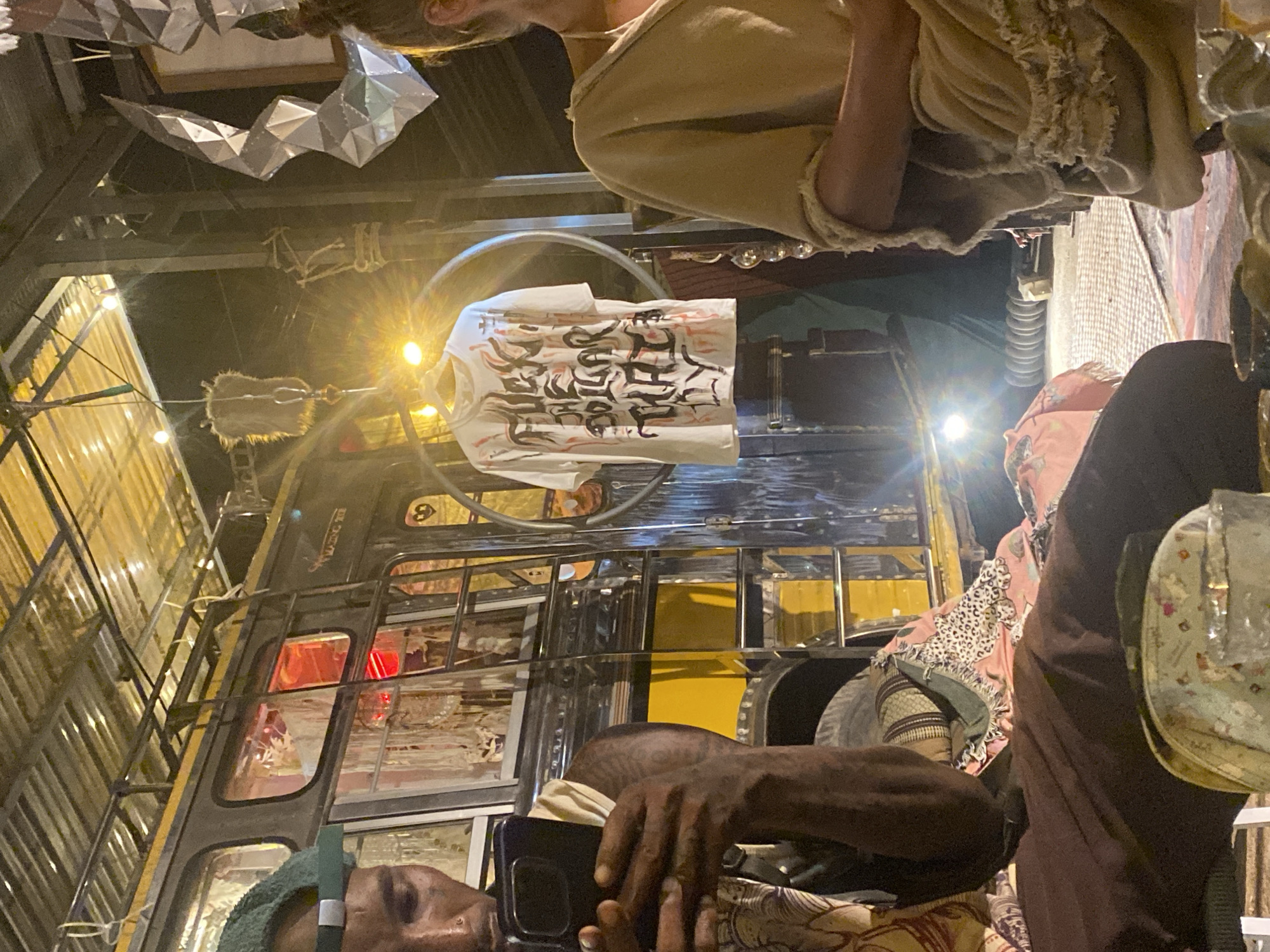 The image, taken in Moo 4, Thailand, shows an unusual composition with the camera tilted upwards. A group of people are visible, with some individuals appearing closer to the lens than others. A person with a green head covering is looking at a phone. A t-shirt with prominent, stylized text hangs in the foreground, suspended from a circular support above a vehicle. The vehicle itself appears to be a converted bus, with yellow and black coloring, and modifications for what looks to be a unique dining or gathering space. The background suggests an outdoor or semi-outdoor venue, with corrugated metal roofing and decorative lighting. The overall ambiance hints at a festive or creative environment.