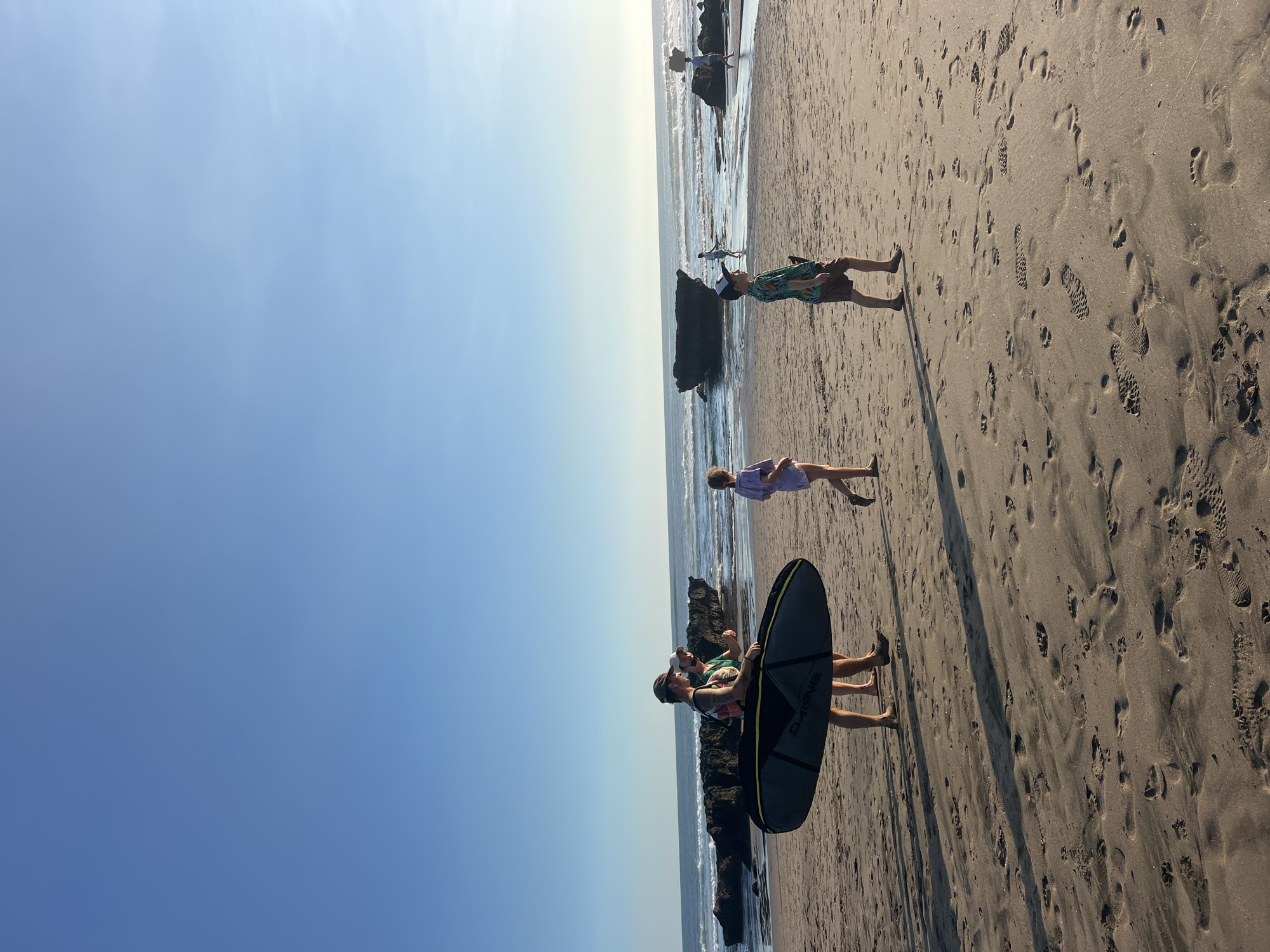 Two adults walk along a sandy beach in North Kuta, Indonesia, carrying a surfboard in a black and yellow bag. To their right, two children walk in the same direction. The ocean stretches out behind them with waves crashing on a rocky shoreline. The sky is clear and blue. The beach is marked with numerous footprints in the sand.
