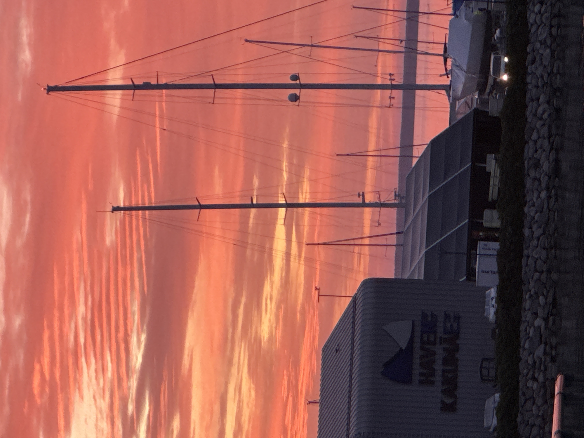 The image shows a marina with sailboats docked, set against a vibrant sunset sky in shades of orange and pink. In the foreground, a building with the text "HAVETS KAKUMÄE" is visible, indicating the location is Kakumäe Harbour in Tallinn, Estonia. The harbour is lined with a stone wall and a grassy area. Several sailboats, with their masts reaching towards the sky, are moored in the water. One sailboat is covered with a tarp. A white car with its headlights on is visible near the boats. The overall scene depicts a tranquil evening at the harbour, illuminated by the warm glow of the sunset.