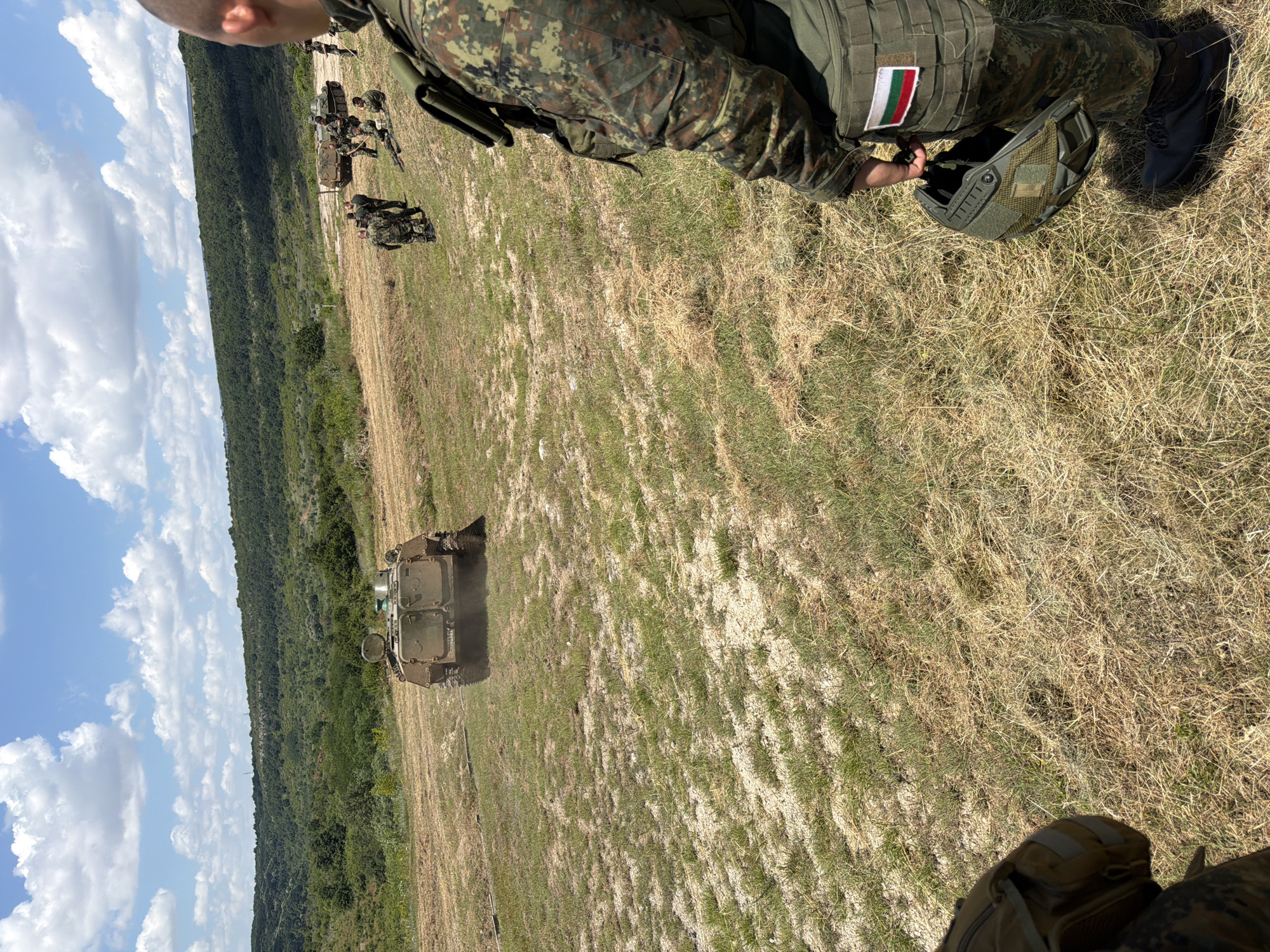 In Beliakovets, Bulgaria, on a sunny day with scattered clouds, a military training exercise is underway. The foreground is dominated by dry, light-colored grass and patches of gravel. A soldier in camouflage uniform, facing away from the camera, is holding a military helmet in their right hand. The soldier's gear includes a tactical vest with a Bulgarian flag patch on the right side. In the middle ground, an armored personnel carrier is driving on a dirt track, kicking up dust. Several other soldiers in camouflage are scattered around, some standing and observing, while others are seated. The background features a lush, green, tree-covered hillside under a bright blue sky.