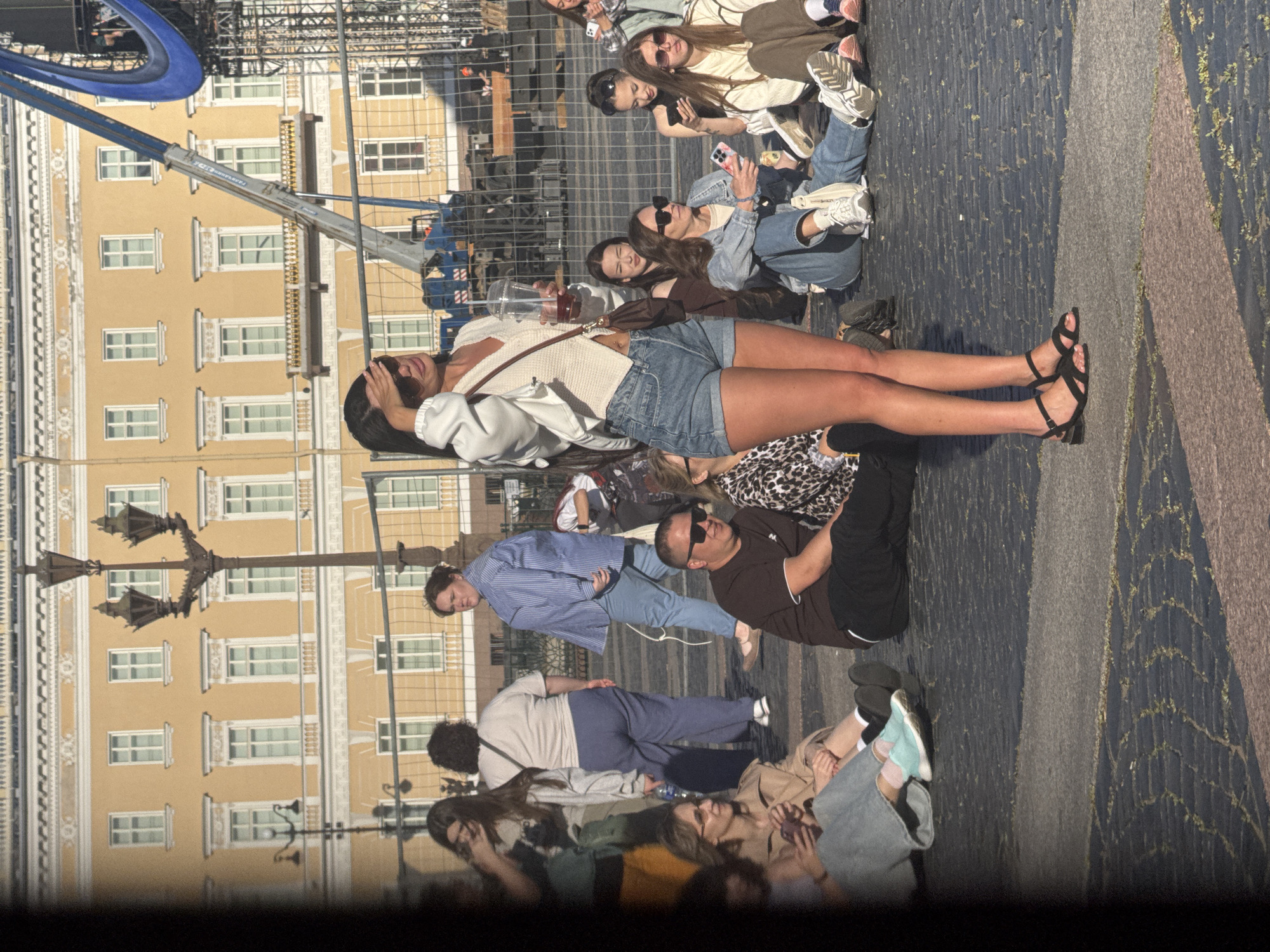 The image depicts a street scene in Saint Petersburg, Russia, likely on a sunny day, with people gathered in an outdoor public space. In the foreground, a young woman is standing, shielding her eyes from the sun with her hand. She is wearing a white open cardigan over a cream-colored top, denim shorts, and black sandals. She holds a clear plastic cup with a beverage. Several other individuals are seated on the ground in the mid-ground, facing various directions. One person in the mid-ground is wearing a denim jacket and jeans, holding a phone. Another is in a white top and brown pants, looking to the side. A man wearing a dark t-shirt and sunglasses is seated closer to the foreground. In the background, a grand, light-yellow building with numerous windows and ornate detailing stands prominently. A street lamp with multiple light fixtures is positioned in front of the building. Construction equipment, including a crane and scaffolding, is visible behind a temporary fence, suggesting ongoing work or an event. The ground is paved with cobblestones. The overall atmosphere appears casual, with people relaxing in an urban setting.