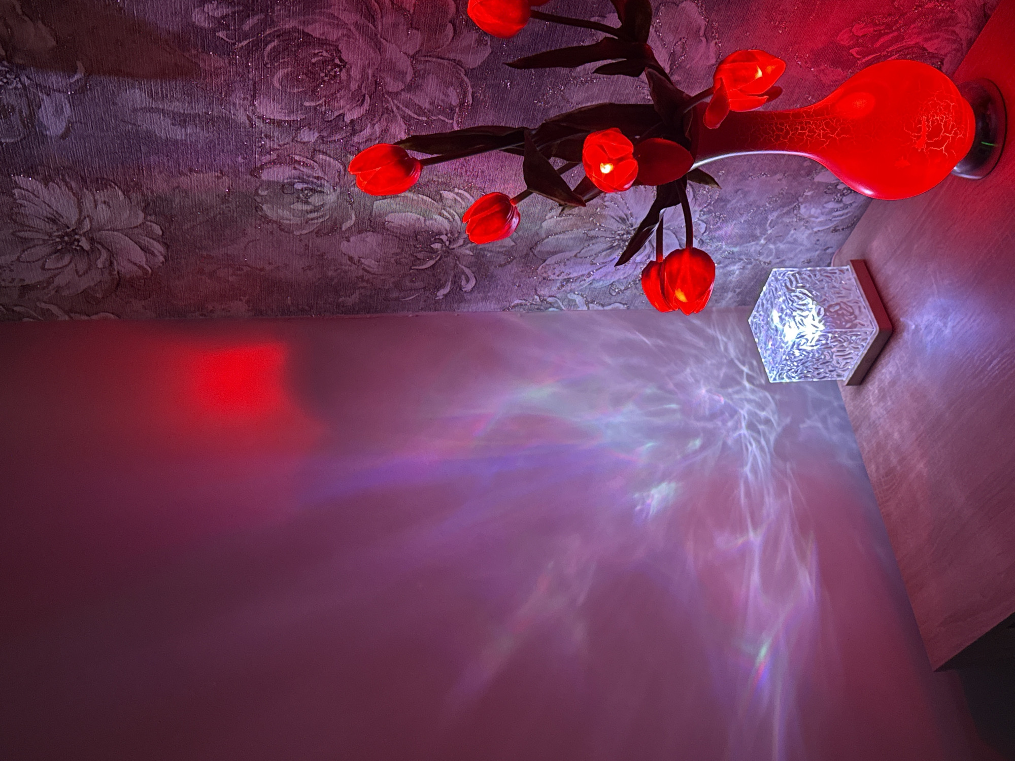 The image depicts an interior view of a room. A wall with a pink hue takes up a significant portion of the lower left. Light patterns create a luminous and ethereal effect. Above, the ceiling is adorned with floral-patterned wallpaper. In the upper right corner is a red vase with tulip-shaped lights emerging from it, mounted on the wall. Next to it, is a hexagonal wall lamp emitting a bright light.