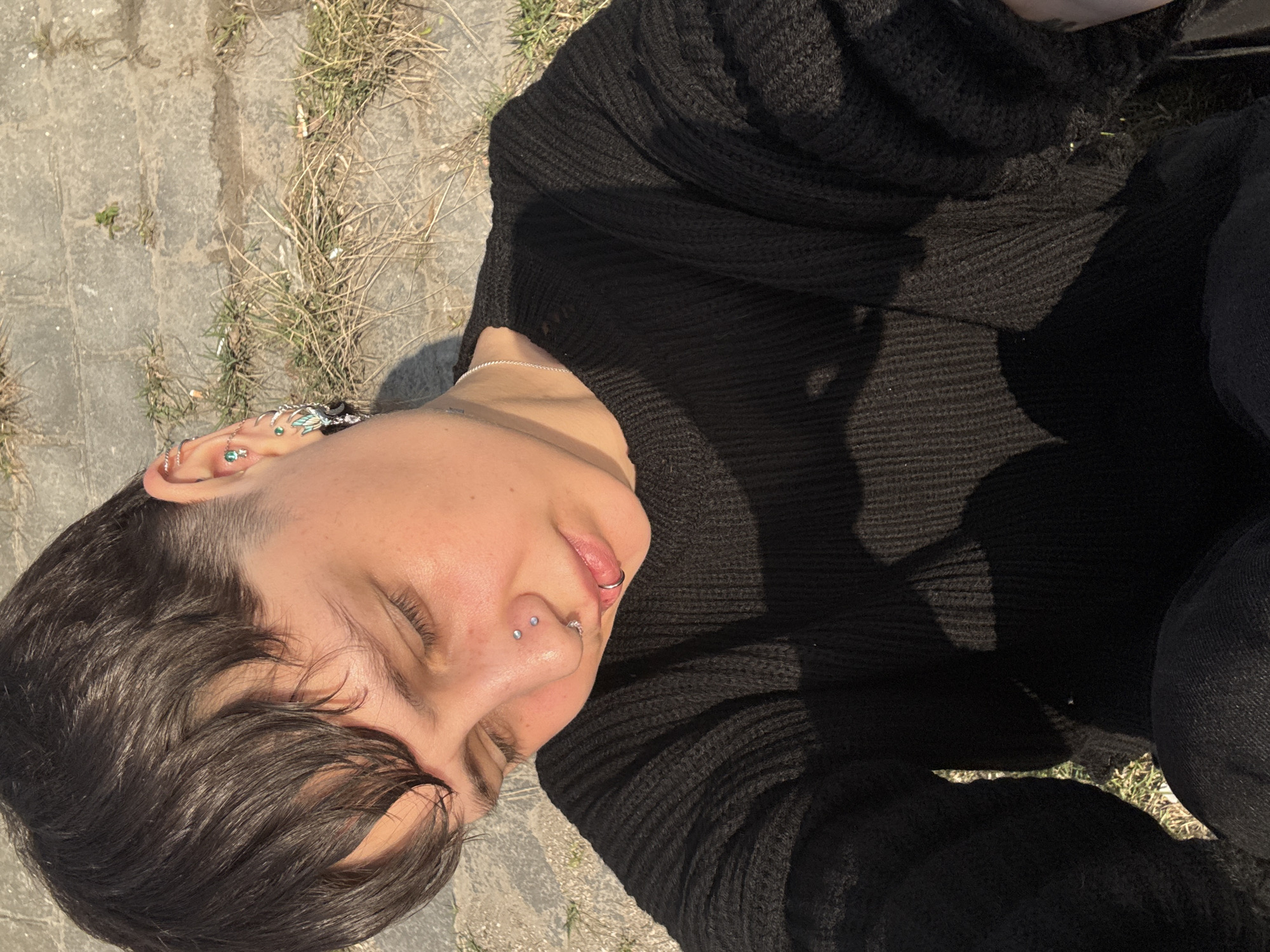 An individual with short, dark hair is lying on their back on a stone or concrete surface adjacent to patches of dry grass and debris. The person is wearing a dark, chunky knit sweater and dark jeans. The individual has multiple piercings in their ear, nose, and lip. The scene is outdoors with bright sunlight. The exact location in Saint Petersburg, Russia, cannot be determined from the image.