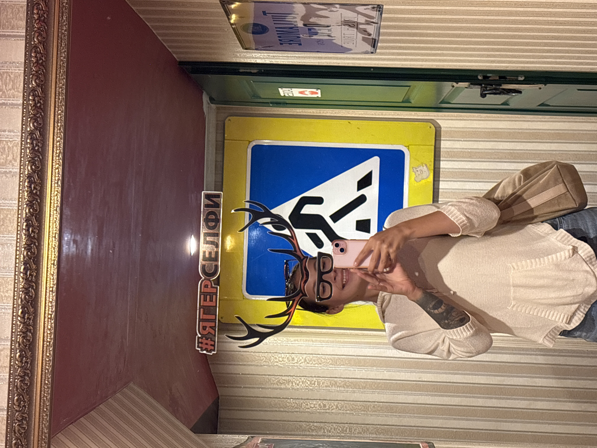 The image is a selfie taken by a person with deer antlers and glasses props, reflecting in a mirror. The person is wearing a beige hooded sweater and jeans, and is holding a pink smartphone. Behind the person, there is a yellow and blue sign with a pedestrian crossing symbol. The environment appears to be indoors, possibly in a bar or restaurant, with striped wallpaper and a decorative ornate mirror frame. There is also a green door and a sign with text on the right side of the image. The hashtag "#ЯГЕРСЕЛФИ" (YagerSelfie) is visible in the mirror. The location is identified as Saint Petersburg, Russia.