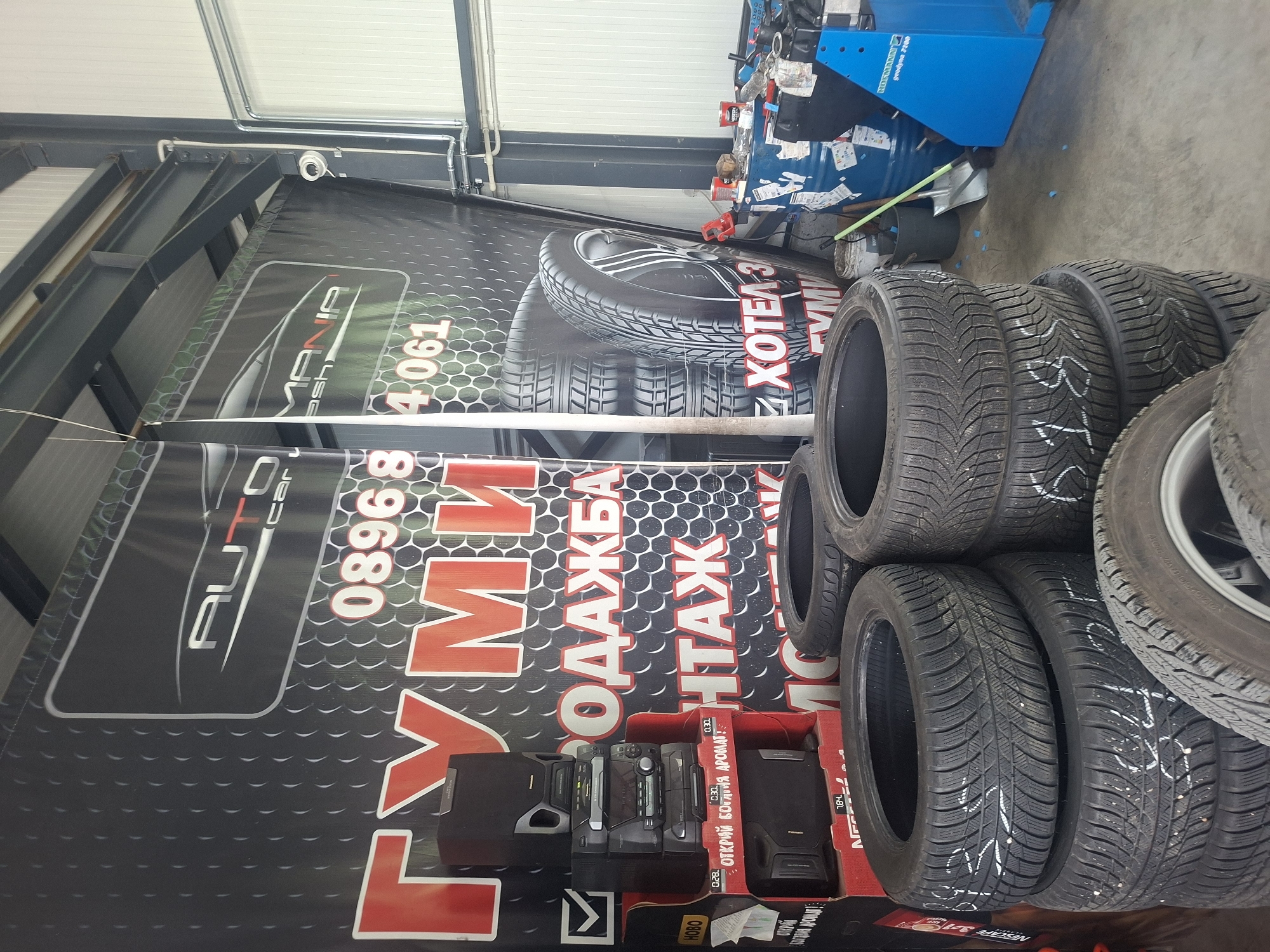 The image shows the interior of a tire shop. A large banner with the words "AUTO CAR MANIA" and a phone number "089684061" is visible. Below the banner are several audio devices in boxes. Stacked tires are seen to the right, some with white markings. A blue tire-changing machine is present in the background. The shop appears to be located in Bulgaria, as there is Cyrillic text on the banner: "ГУМИ ПРОДАЖБА МОНТАЖ" and a product advertised with "ОТКРИЙ БОЛГИЯ АРОМАТ".