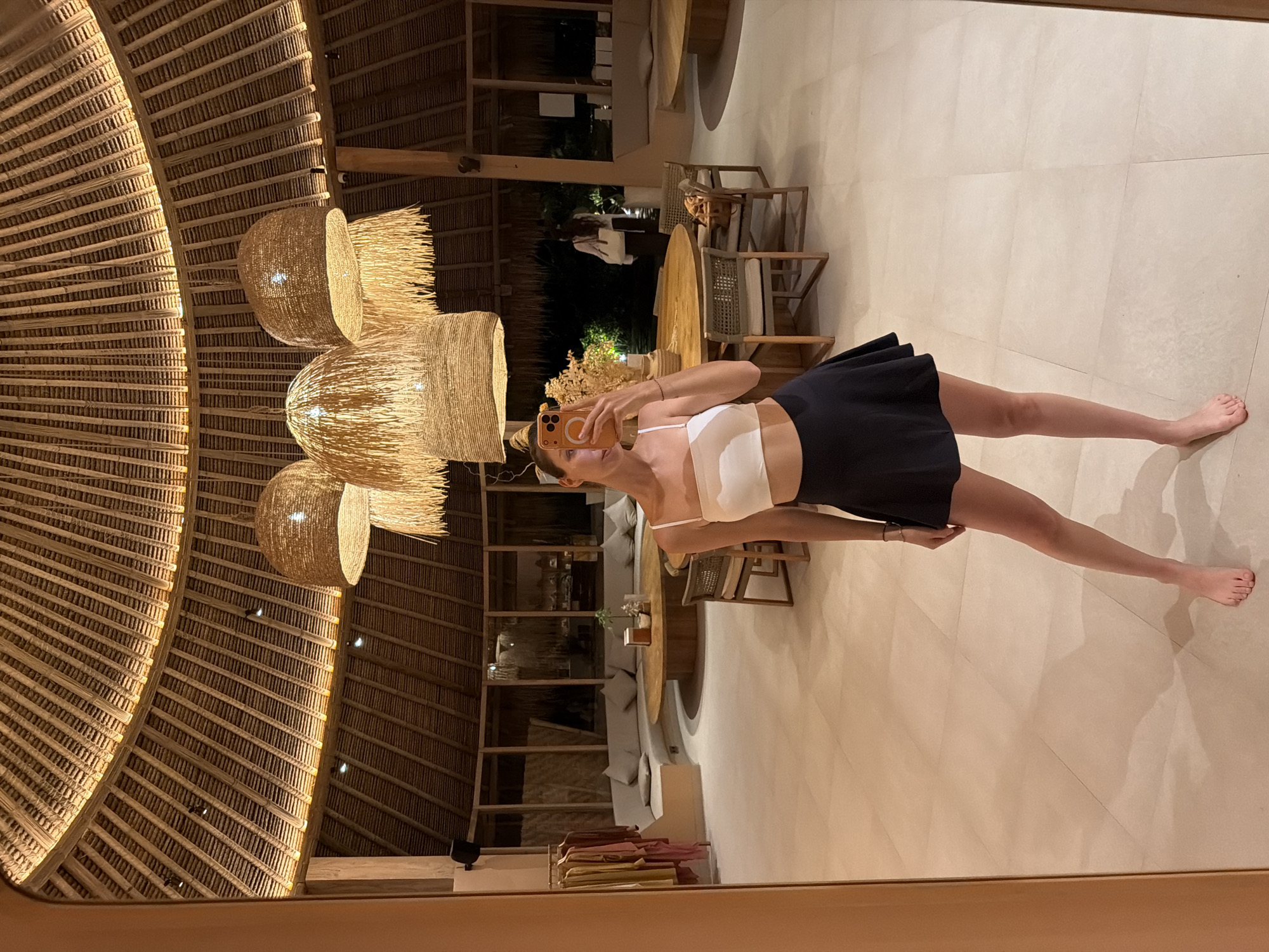 A female subject is depicted taking a mirror selfie indoors. She stands barefoot, facing the mirror directly, holding an orange-cased smartphone in her right hand, obscuring her face. She wears a white crop top with thin straps and a dark, pleated mini-skirt. Her hair is pulled back, secured with a light-colored accessory. A light-colored bracelet is visible on her right wrist.
The setting is a spacious, circular room with a high, domed ceiling constructed from radially arranged natural wood or thatch. Multiple large, woven natural fiber pendant lamps hang centrally, emitting warm light. Recessed spotlights illuminate the ceiling's perimeter. The floor is covered with large, light-colored square tiles.
In the background, a large, curved, light-grey upholstered sectional sofa with throw pillows is arranged around a circular wooden coffee table, on which a small plant arrangement sits. Further back, a larger circular wooden dining table is surrounded by chairs featuring woven backs and light-colored cushions. On the far left, a clothing rack displays various garments. Through large windows or open archways behind the furniture, a dark exterior is visible, indicating nighttime. A faint reflection of another person, a female walking away, is visible near the windows, wearing a white top and carrying a light-colored bag. The structure incorporates natural wood pillars and a light-toned interior palette. The location is in the city of Unknown, Indonesia.
