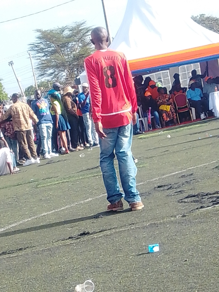 A man in a red "8 AS HÜRTH" jersey and jeans stands on a green field scattered with litter. He is facing away from the camera, towards a crowd of people and a tent-like structure. The crowd is varied in attire, some sitting on chairs under the tent. Trees are visible in the background under a clear sky. The lighting is bright, casting shadows on the field.