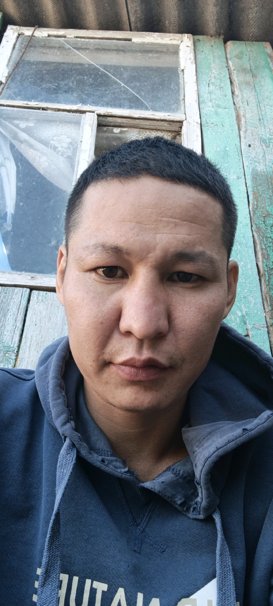 A close-up, frontal, eye-level, waist-up shot shows a person with short, dark hair and brown eyes. The individual is wearing a dark blue hooded sweatshirt. The background features a dilapidated window with a cracked pane and peeling green paint on wooden planks. The scene suggests an outdoor, possibly rural, setting. The overall impression is one of wear and tear, possibly indicative of a rural dwelling or abandoned structure. The exact location within Раздор, Russia cannot be determined from the image alone.