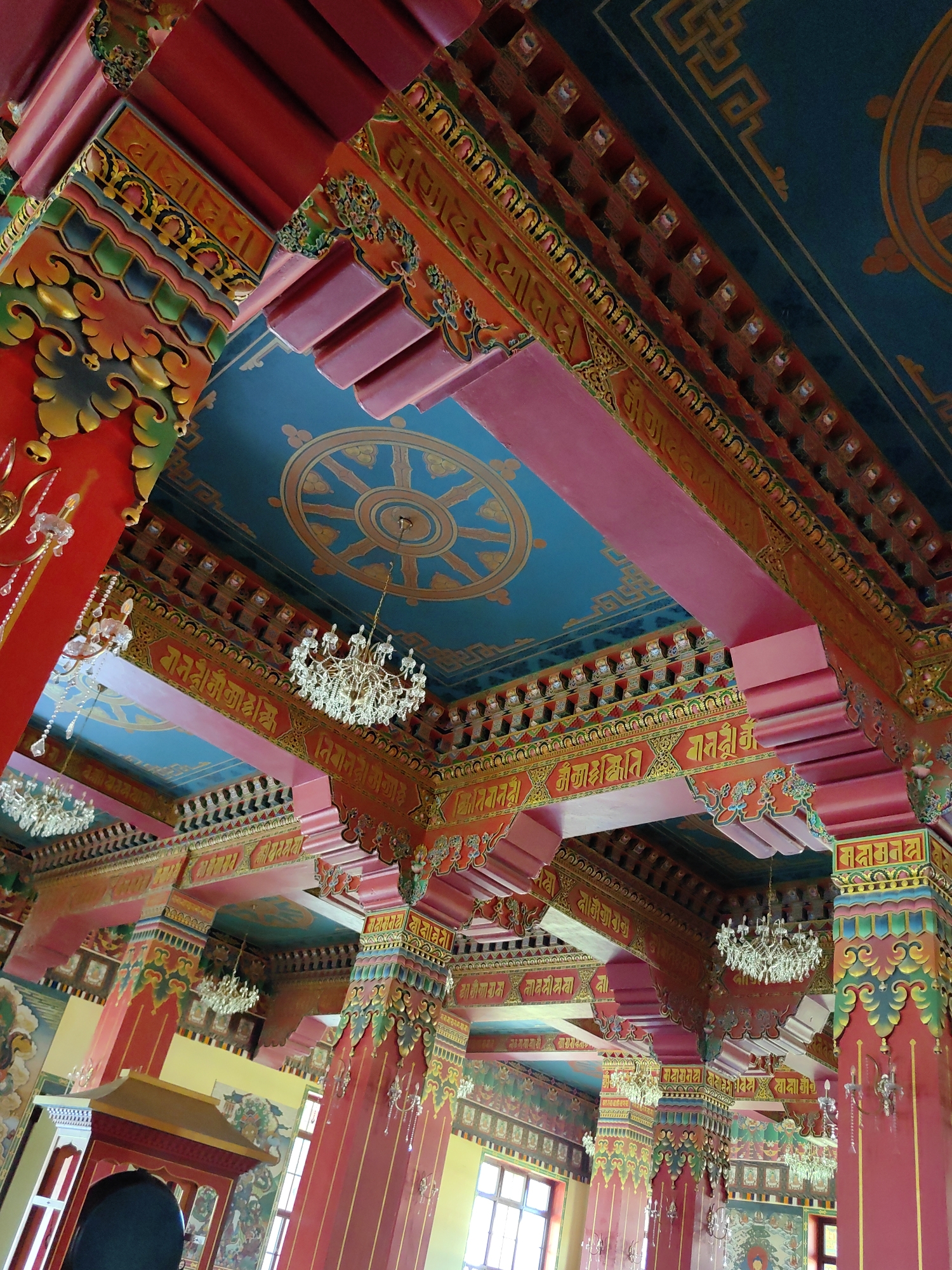 The image shows the interior of a temple, likely in Parsa, Nepal, characterized by vibrant colors and intricate architectural details. The ceiling is painted a deep teal blue, featuring a prominent golden Dharma wheel. Ornate red and orange beams, adorned with decorative patterns and what appear to be Tibetan script, support the ceiling. Several crystal chandeliers hang from the ceiling, illuminating the space.

Large, red columns, decorated with colorful motifs in green, yellow, and blue, rise from the floor, supporting the upper structures. These columns are also embellished with decorative carvings and possibly further script. In the background, sections of murals or paintings are visible on the walls, depicting figures and scenes, adding to the temple's rich visual tapestry. The overall impression is one of opulence and spiritual significance, with a strong emphasis on traditional Buddhist art and architecture. There are no people clearly visible in the frame.