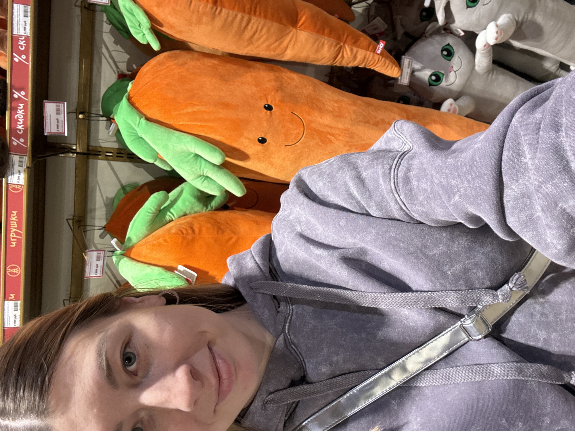 A vertically oriented medium shot depicts a woman in a gray hoodie, partially obscuring stuffed carrot toys in a store. The orange carrot toys are displayed on a shelf. A red promotional sign labeled "скидки %" is visible on the left. Other stuffed toys, possibly cats, are present on the shelf. The setting is potentially in a retail store in Moscow, Russia, based on the Cyrillic text on the promotional sign.