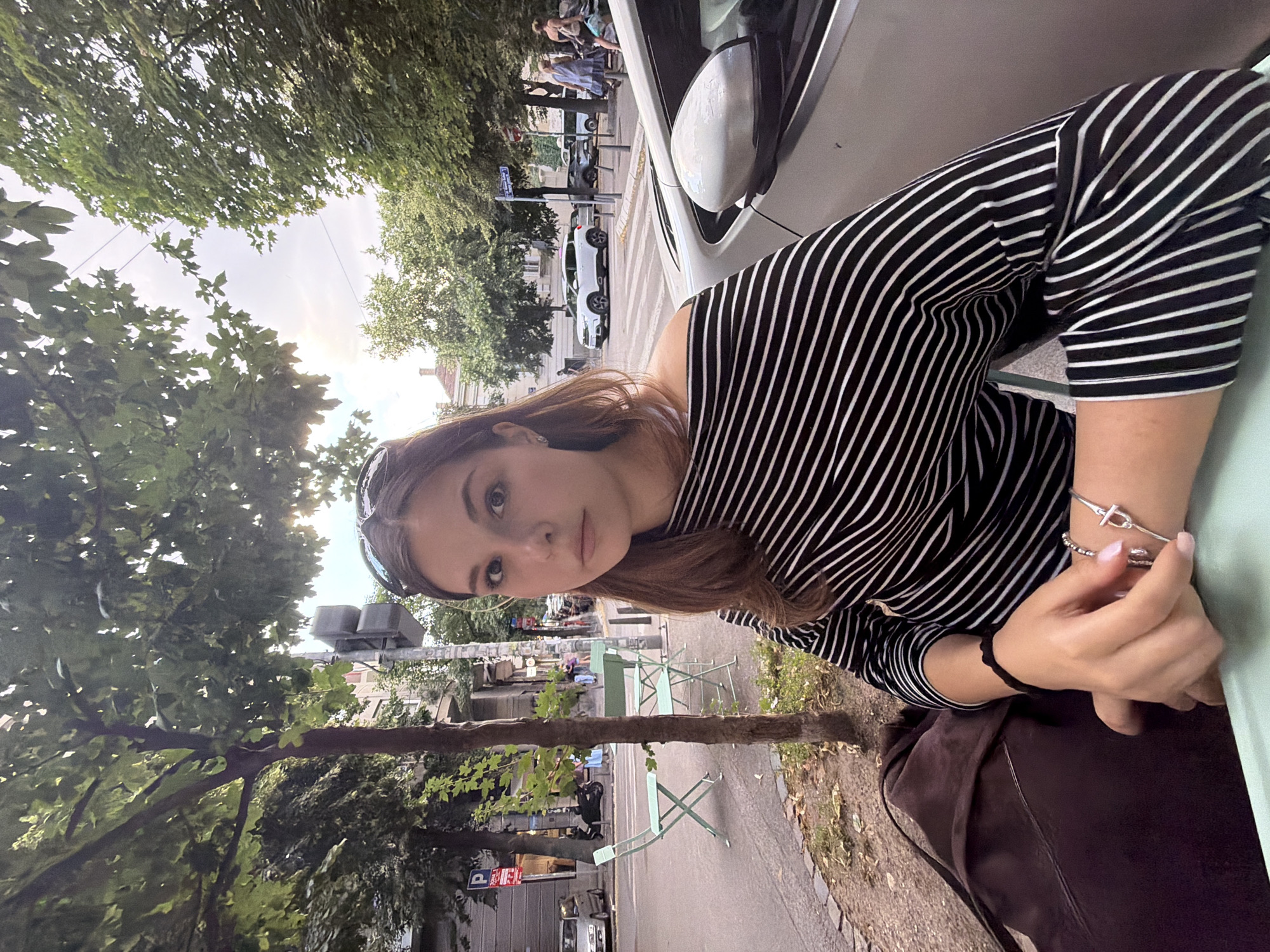 A young woman with long brown hair is pictured in a medium shot, seated outdoors at a cafe in Belgrade, Serbia. She is wearing a black and white striped off-the-shoulder top. Her hands are resting on a light green table. On her right wrist, she wears a silver bracelet. To her left, a dark brown bag is visible on her lap. The background reveals a street scene with parked cars, a tree, and buildings. Green metal chairs and tables are set up on the sidewalk. The lighting suggests it is daytime, with sunlight filtering through the leaves of the tree.