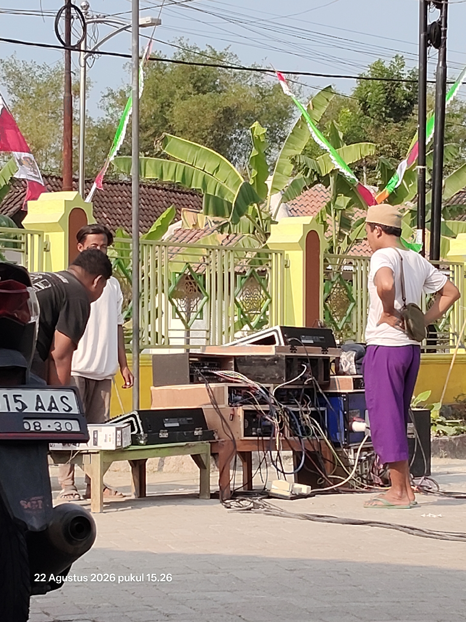 In Wedi, Indonesia, on August 22, 2026, at 3:26 PM, a street scene unfolds. The foreground is dominated by the rear of a dark motorcycle with a visible license plate reading "15 AAS 08-30". To the right, a man wearing a white t-shirt and purple pants stands by a complex setup of audio equipment. He has a small green bag slung over his shoulder and is facing away from the camera, with his hands on his hips. Behind him, another man in a black t-shirt is bending over, seemingly working on or inspecting the equipment. A third person in a white t-shirt is standing between them. The background features a yellow building with a green fence, adorned with colorful flags that are characteristic of local celebrations. Banana trees with large green leaves are visible behind the fence, suggesting a tropical or subtropical environment. Power lines crisscross the sky, and the ground is paved with light-colored interlocking bricks. The overall atmosphere appears to be one of preparation or setup for an outdoor event.