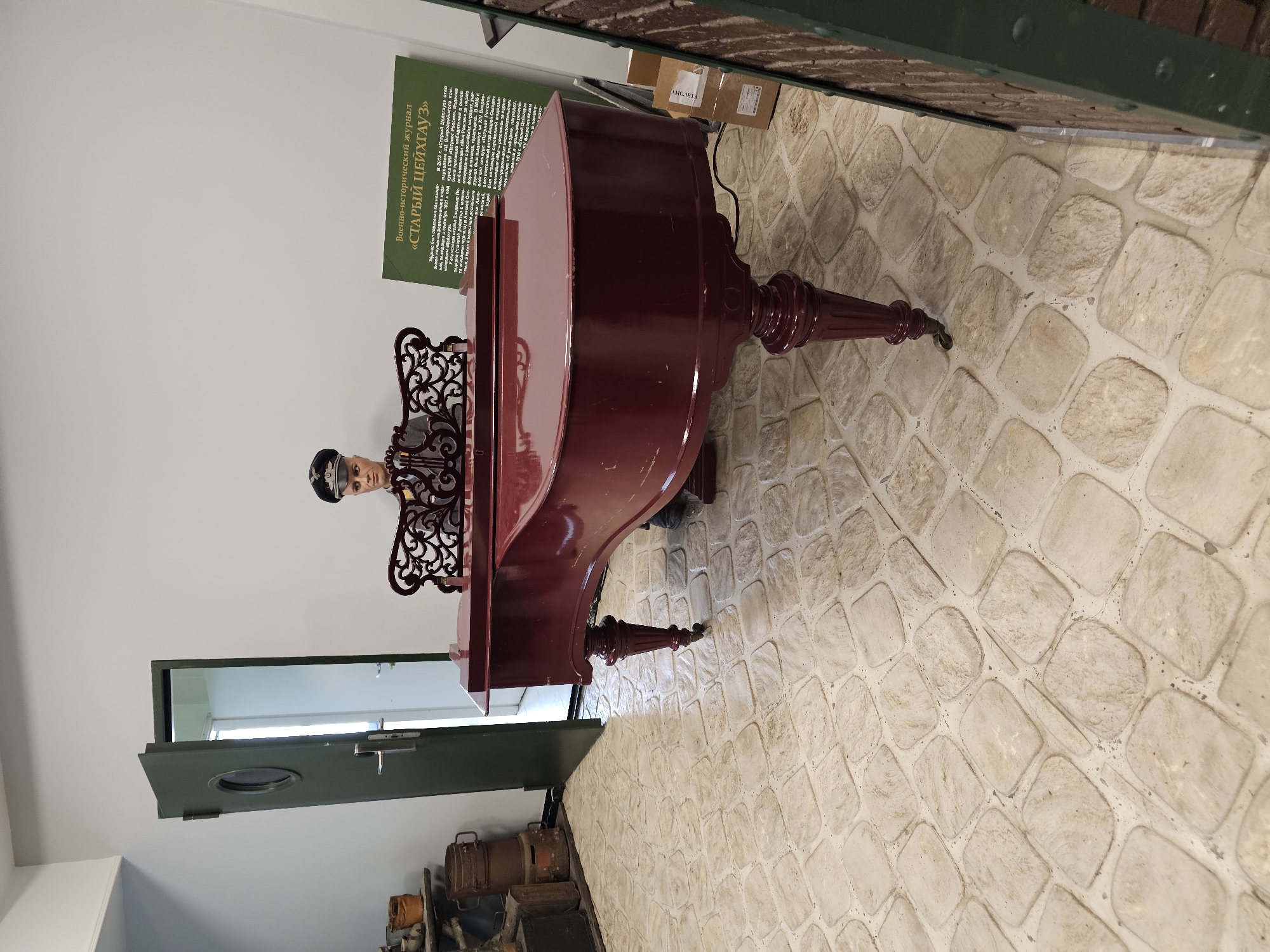 The image captures a scene within a building, potentially a museum or historical site, possibly in Архангельское, Russia. A reddish-brown grand piano dominates the scene, placed on a floor with light-colored stone tiles. Behind the piano is a lifelike mannequin dressed in what appears to be a historical military or official uniform, positioned as if playing the instrument.
A green sign with Russian text is mounted on the wall near the piano, likely offering information or context about the display. To the left, a partially open dark green door leads to another room with a glimpse of a white interior and window. Below the doorway, a collection of vintage suitcases and trunks are arranged.
The room's decor and the presence of the mannequin and informational sign suggest a staged exhibit or historical recreation.
