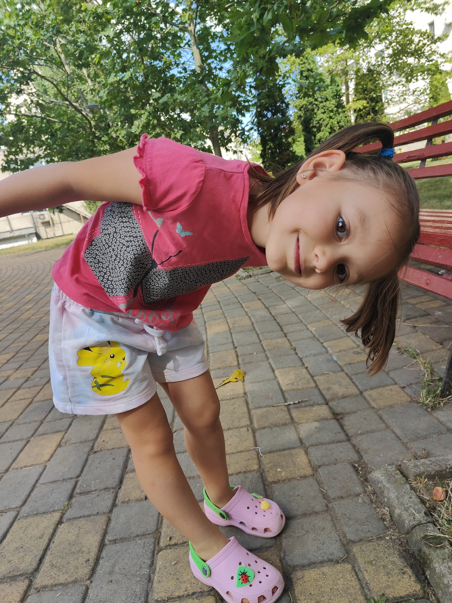 A young girl with brown hair tied in pigtails wearing a pink t-shirt and white shorts with a Pikachu design on them is crouching down in a park. She is wearing pink Crocs with green accents and a ladybug decoration. The girl is looking towards the camera with a slight smile. Her t-shirt has a graphic design on it.
The setting appears to be an outdoor park with a paved walkway made of interlocking stones. There are trees with green foliage and a red park bench visible in the background. The lighting suggests it is daytime, likely with sunny weather. The scene is set in Chornomorsk, Ukraine. There is no visible text in the image.