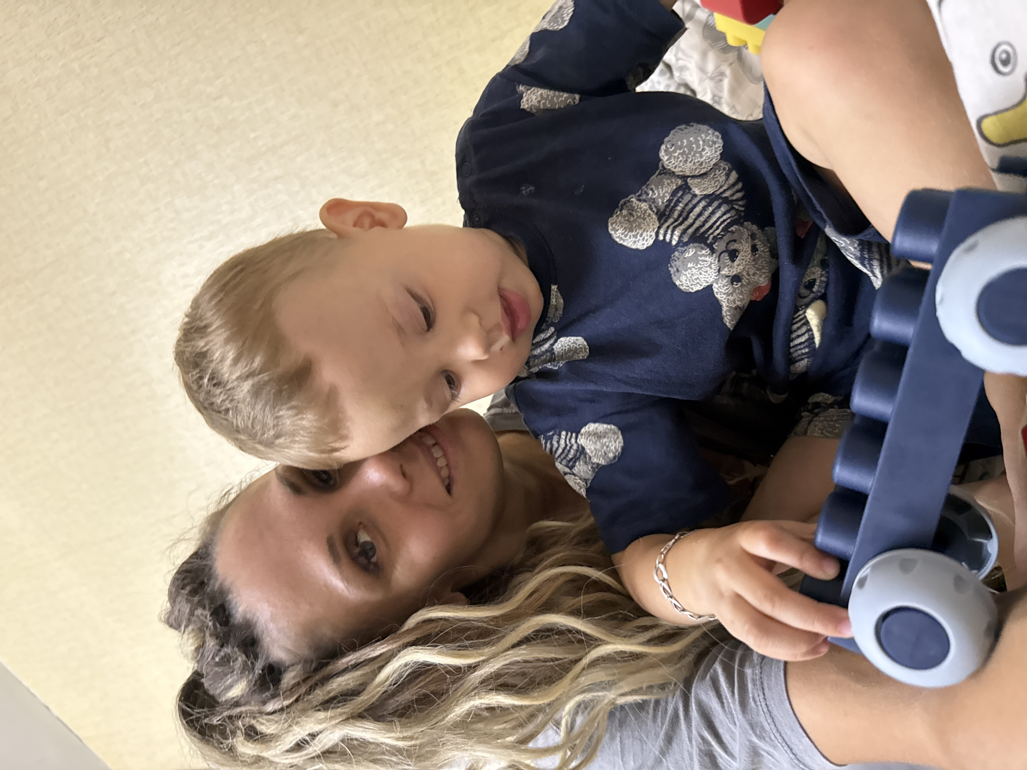 A woman with blonde, curly hair pulled back in a ponytail and wearing a grey t-shirt is seen from the chest up, looking towards the camera and smiling. Beside her, a young child with short blonde hair, wearing a navy blue t-shirt with a pattern of teddy bears, is looking down. The child is holding a blue toy with grey wheels. The background is a beige textured wall, suggesting an indoor setting, possibly a home. The image appears to have been taken in Rusanovo, Russia.