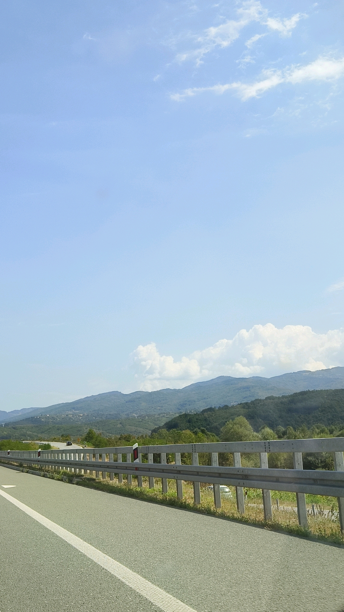 The image depicts a view from a highway in or near Vranje, Serbia, looking towards a mountainous landscape under a bright blue sky with scattered white clouds. In the foreground, there is a paved road with a white line marking the lane. A metal guardrail runs alongside the road, separating it from the embankment. Beyond the guardrail, there is green vegetation and some distant trees. The mountains in the background are covered in a mix of trees and appear to be rolling hills. The overall scene suggests a sunny day, likely during daylight hours. There are no visible people in the frame, and the only detected vehicles are a couple of cars in the distance, one on the highway and another parked off to the side.