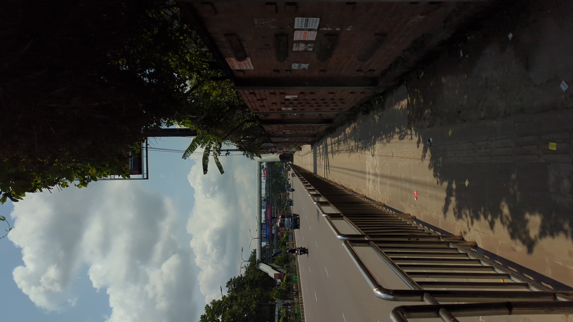 A perspective view of a busy Dhaka, Bangladesh street taken from an elevated pedestrian overpass. The scene is dominated by a wide road teeming with vehicles. The lower portion of the image shows the elevated overpass's metal railings. To the left of the road, lush green trees and vegetation create a contrast to the urban environment. Above this greenery, a blue sky is scattered with puffy white clouds.
On the right side of the image, a brick wall runs along the side of the overpass, its surface marked with patches of peeling posters and shadows. Further down the road, a tunnel or overpass is seen, providing a glimpse of the cityscape beyond. Streetlights line the sides of the road, adding to the urban feel. Vehicles, including cars and motorcycles, move along the road.