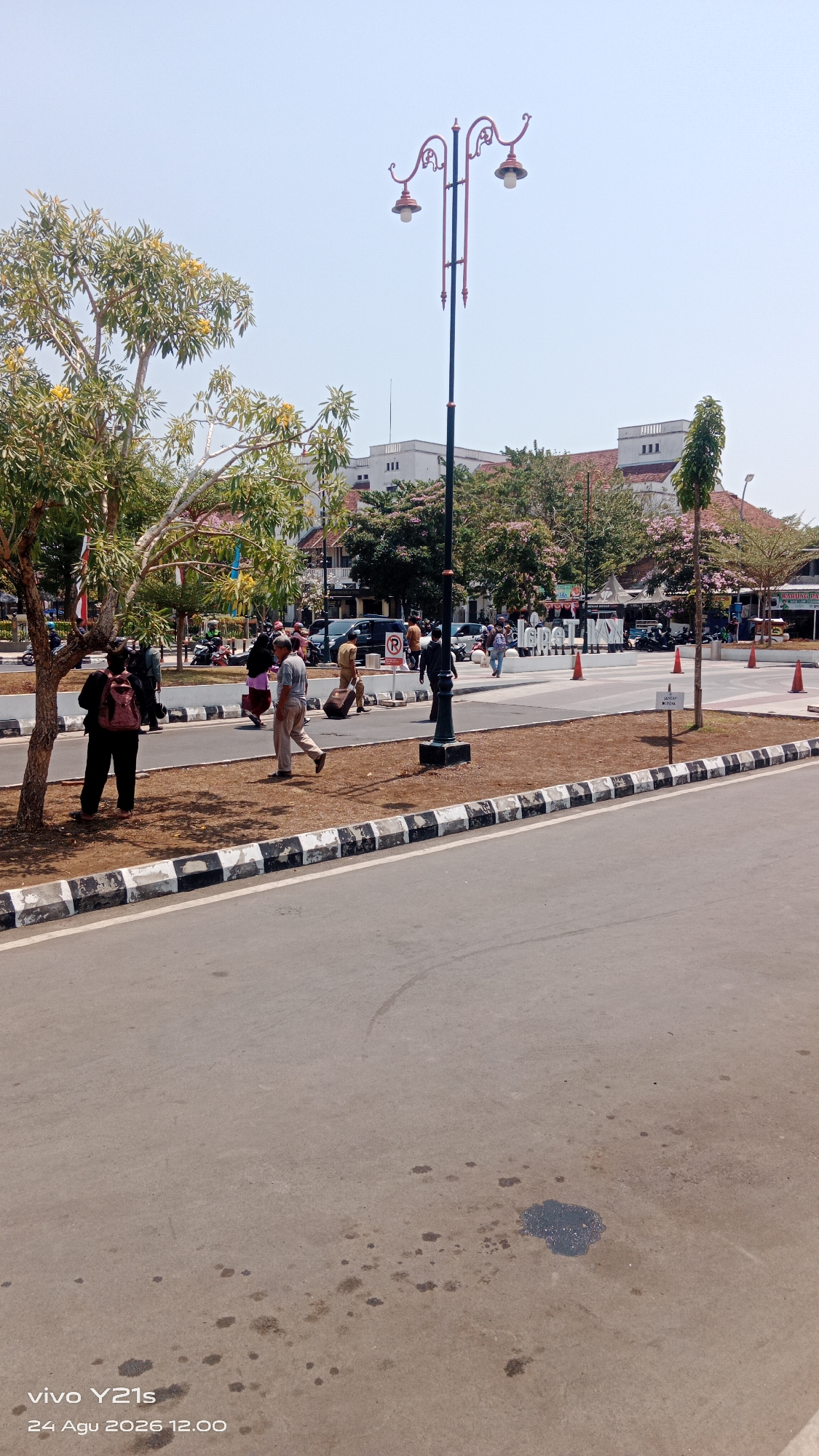 The image depicts a street scene in Tegal, Indonesia, likely in a public square or pedestrian area. In the foreground, a paved road with oil stains is visible. Adjacent to the road is a median strip with grass and decorative black and white patterned curbing. A streetlamp with an ornate design featuring two lamps stands in the middle ground, casting no light as it is daytime. Several people are walking or standing on a paved area behind the median. One man is pulling a suitcase, while others appear to be conversing or walking. A large tree with yellow flowers is on the left side of the image, partially obscuring a view of buildings in the background. The buildings are light-colored with red roofs and appear to be commercial or institutional structures. In the far background, traffic is visible on a wider road, and a sign with the letters "lsp LKK" is present. The sky is clear and bright, suggesting a sunny day. The image is watermarked with "vivo Y21s 24 Agu 2026 12.00".
