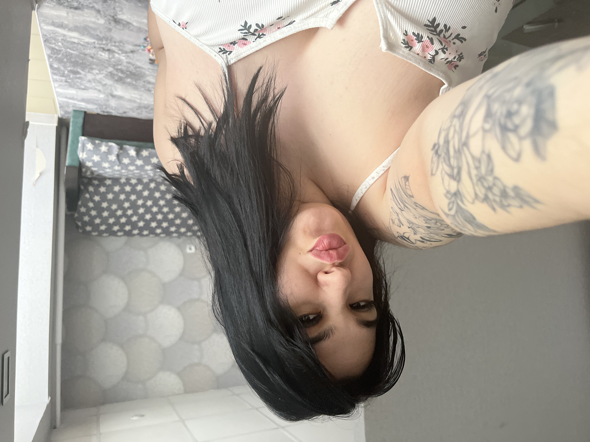 A woman with long black hair is looking at the camera. She is wearing a floral top. Her right arm is visible and has a tattoo. The location is possibly a bathroom or bedroom. A bed is visible in the background. The image was taken indoors. There is no indication of a location in the city of Chaikovsky, Russia.