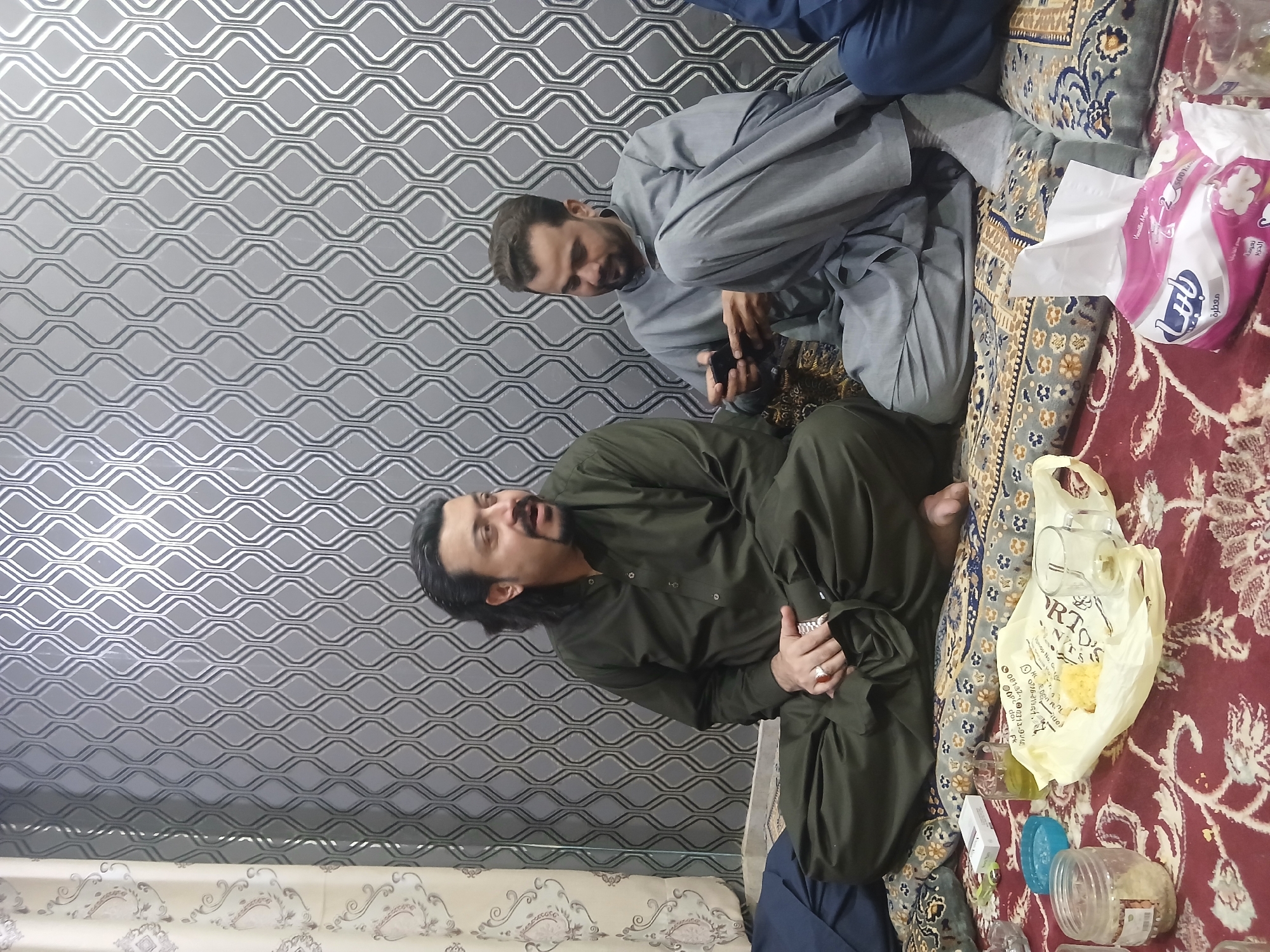 Two men are sitting on a patterned cushion on top of a red patterned rug, seemingly indoors. The person on the left is wearing a dark green shalwar kameez, while the person on the right is in a light gray shalwar kameez and looking at a phone. On the rug are a box of tissues, a yellow bag with what appears to be food inside, and a few plastic containers. The background features a wavy gray patterned wall. The location is Quetta City Tehsil, Pakistan.