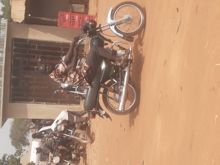 The scene depicts an unpaved, reddish-brown dirt road in the city of Gwadanye, Nigeria, under clear daylight. In the foreground, a man is seated on a dark-colored motorcycle, facing slightly left, gripping the handlebars. He wears a multi-patterned short-sleeved shirt, dark trousers, and sandals. The motorcycle features a chrome exhaust pipe, a silver front fender and wheel, and a dark rear wheel.
In the midground to the left, at least two additional motorcycles are present. One man in a striped shirt is seated on a motorcycle, while another man in a white shirt is bent over a separate motorcycle, appearing to tend to it. Additional figures are partially obscured further in the background.
The background consists of several structures: a concrete building with a dark, gridded metal door is centrally located. To its right, a simpler building features stacked red Coca-Cola crates outside. On the far left, a portion of a white cylindrical tank, marked with "PK," is visible. Shadows on the ground indicate a sunny environment.