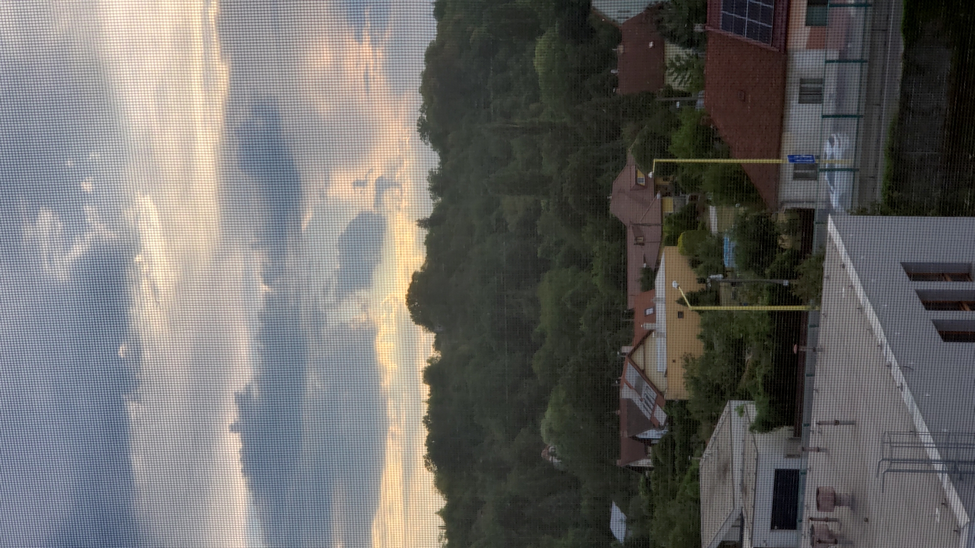 The image depicts a view through a window screen of a residential area with a wooded hill in the background. The sky is cloudy with patches of sunlight. In the foreground, there are several buildings with red-tiled roofs and one building with a yellow facade. A street with a white car is visible. The image appears to be taken from an elevated position. The location is identified as Prešov, Slovakia.