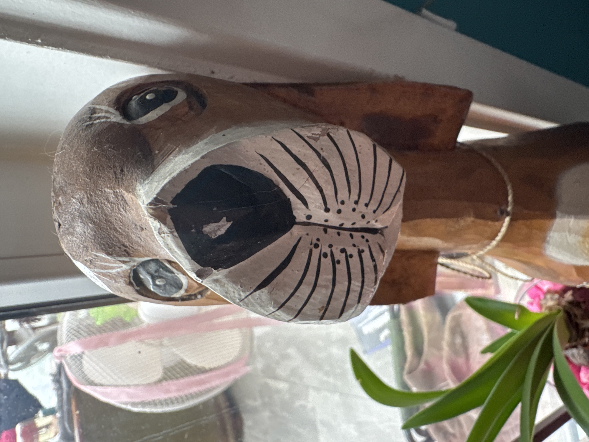 This close-up, vertical photograph depicts an indoor scene in Unknown, Unknown. The central subject is a carved wooden sculpture of a dog's head and upper torso, facing directly towards the viewer. The sculpture is predominantly brown wood, with painted details. The dog's eyes are black with small white highlights, encircled by white paint. Its snout is a prominent white area with a black nose and several thin black lines simulating whiskers, accompanied by small black dots on the cheeks. White lines also extend above the eyes. Flappy ears are carved into the sides of the head. A thin, light-colored cord or rope is loosely tied around the sculpture's neck. In the bottom-left foreground, a green plant with several broad, pointed leaves is partially visible, rooted in a container filled with pink decorative pebbles. The background, visible through a window, displays a blurred outdoor or indoor area. A white oscillating fan with a pink ribbon or band tied around its cage is clearly discernible through the window. A plain white vertical surface is visible to the right of the sculpture, with a dark teal or blue-green surface partially visible further right.