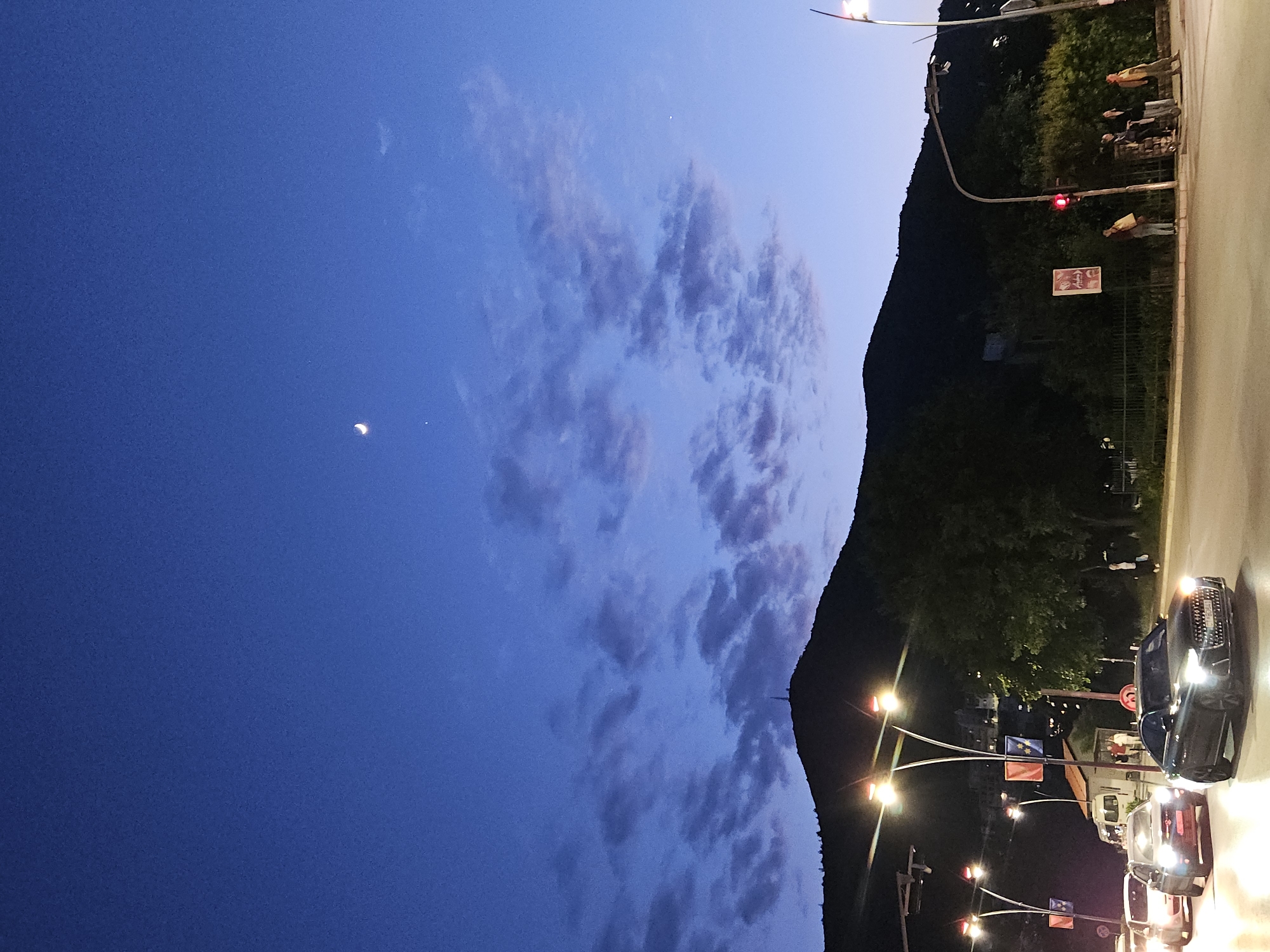 The image depicts an evening scene in a city, likely near Unknown. The sky is a deep blue, with light pink clouds to the left. A crescent moon is visible in the upper left corner. The right side of the image shows a tree-covered mountain and an adjacent street. There are several street lights and parked cars with their headlights on. A traffic light can be seen on the street, and individuals can be seen on the sidewalk.