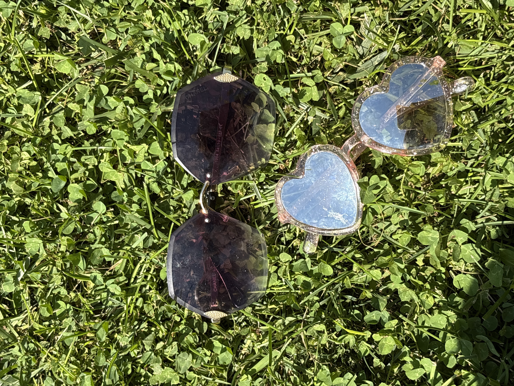 Two pairs of sunglasses are resting on green grass, likely in Oreshak, Bulgaria, under natural sunlight. The upper pair has dark, polygonal lenses with a gold accent on the temples. The lower pair is heart-shaped with light blue lenses and a glittery pink frame. The grass is composed of a mixture of clover and blades of grass.