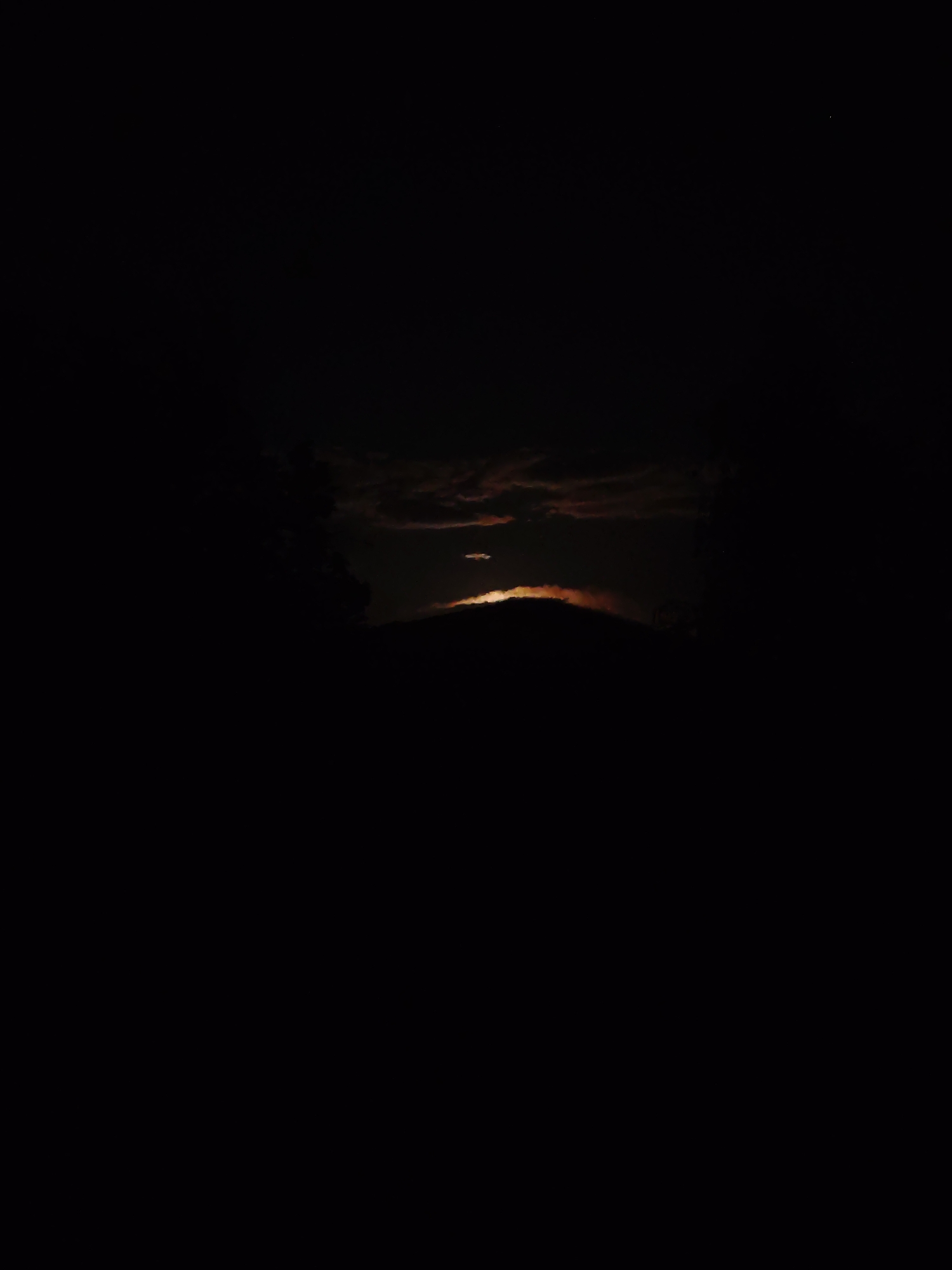 The image presents a striking nocturnal or twilight landscape over Starozagorski bani, Bulgaria, characterized by extreme darkness punctuated by a dramatic natural light phenomenon. At the center, a bright, horizontal band of light, likely the moon or a very low-lying sun, emerges from behind a dark mountain or hill silhouette. This celestial body illuminates the edges of a layer of clouds directly above it, creating vivid streaks of orange and reddish light against the otherwise dark sky. Higher up, other cloud formations are faintly discernible as dark masses.
The foreground and much of the upper sky are rendered in deep black, emphasizing the powerful contrast of the illuminated central elements. To the left, a dark, indistinct mass of what appears to be trees or dense foliage forms a silhouette against the slightly lighter upper background. The overall scene conveys a sense of serene mystery and natural grandeur. There are no people, man-made objects, or visible text within the frame. The time of day is clearly night or the very early/late hours of dawn/dusk, and the weather appears to be partially cloudy.