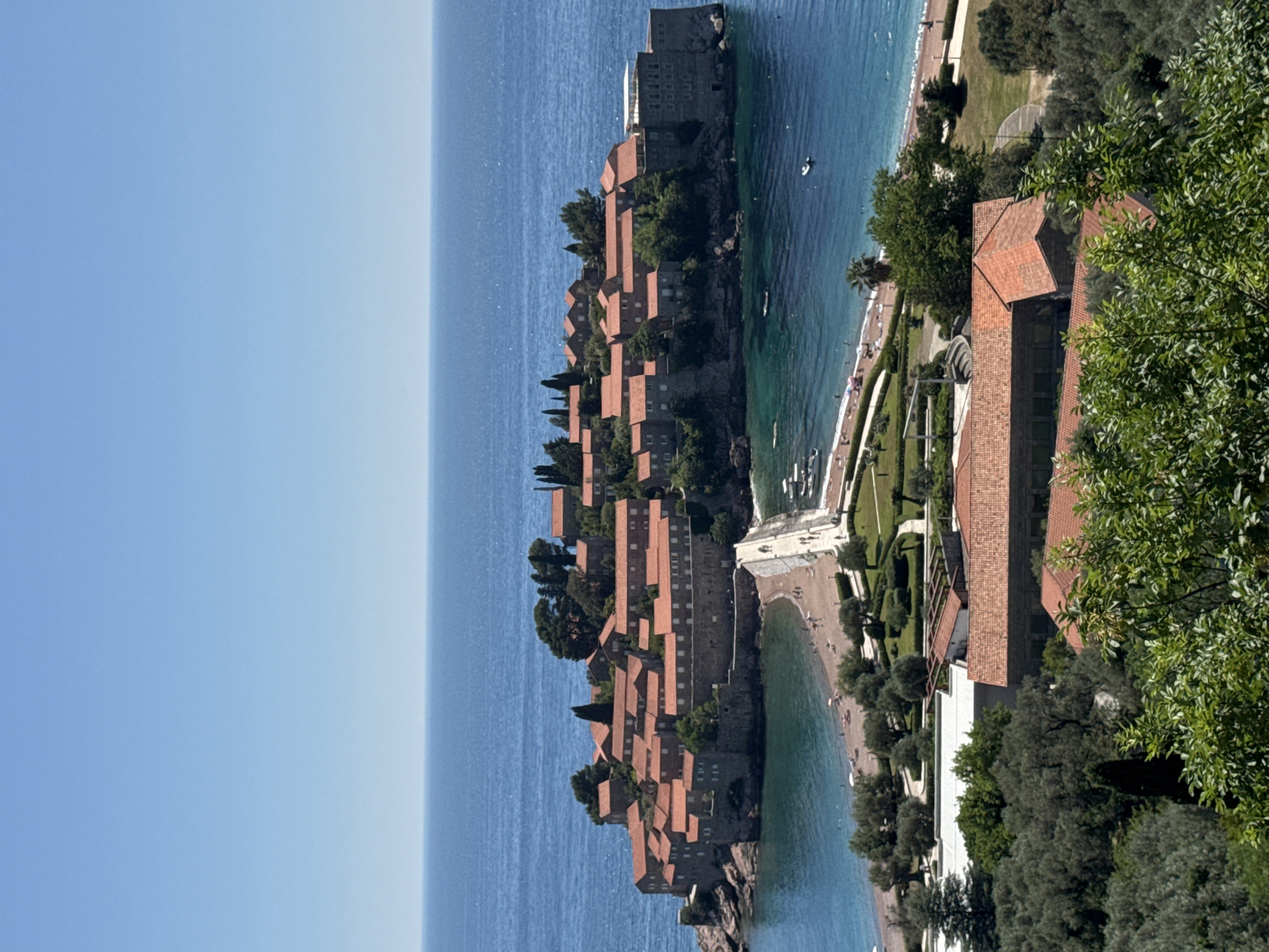 The image depicts Sveti Stefan, Montenegro, a small islet connected to the mainland by a narrow isthmus. The islet is densely packed with buildings featuring terracotta roofs and stone walls, surrounded by lush greenery. The deep blue sea stretches out towards the horizon, meeting a clear blue sky. On the mainland side, there's a beach with people relaxing, along with manicured gardens and more buildings, partly obscured by trees. The scene is brightly lit, suggesting a sunny day.