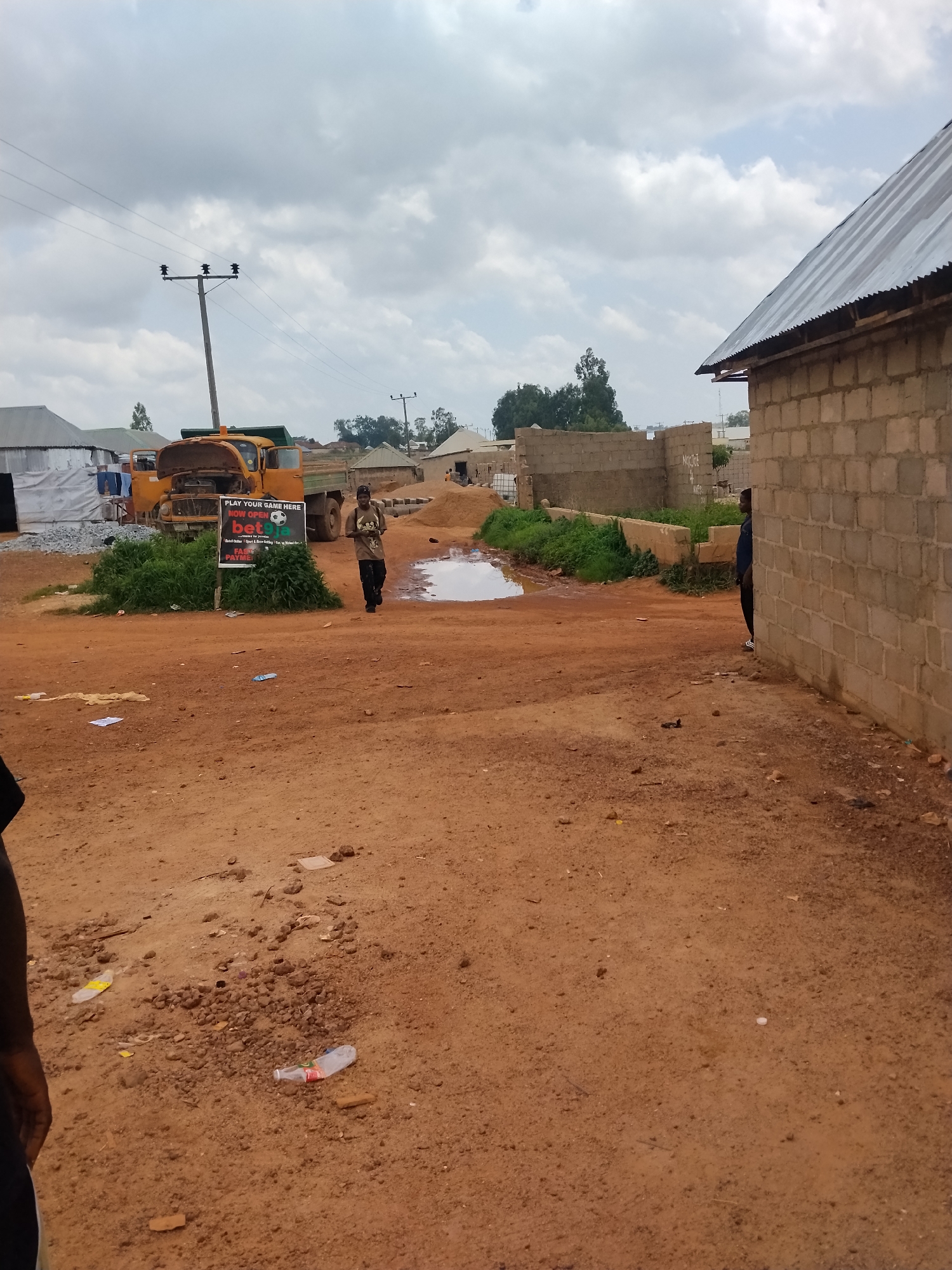 The image depicts an outdoor scene in Durumi, Nigeria, likely a construction or development area. In the foreground, a dirt path is scattered with small stones, dirt, and some litter, including plastic bottles. To the left, a person's arm is visible, suggesting the photographer is present. Further back, a yellow dump truck with its hood up is parked. A sign in front of the truck reads "PLAY YOUR GAME HERE NOW OPEN betgo" and "FAST PAYMENT". A person in a dark shirt and pants is walking away from the sign, towards the right side of the image. To the right, a partially constructed building made of cinder blocks stands, with a corrugated metal roof. Another person in dark clothing is partially visible peeking out from behind the building. In the background, there are more buildings, trees, and utility poles with electrical wires against a cloudy sky. The overall environment appears somewhat arid with patches of green grass and a small puddle of water on the dirt path.