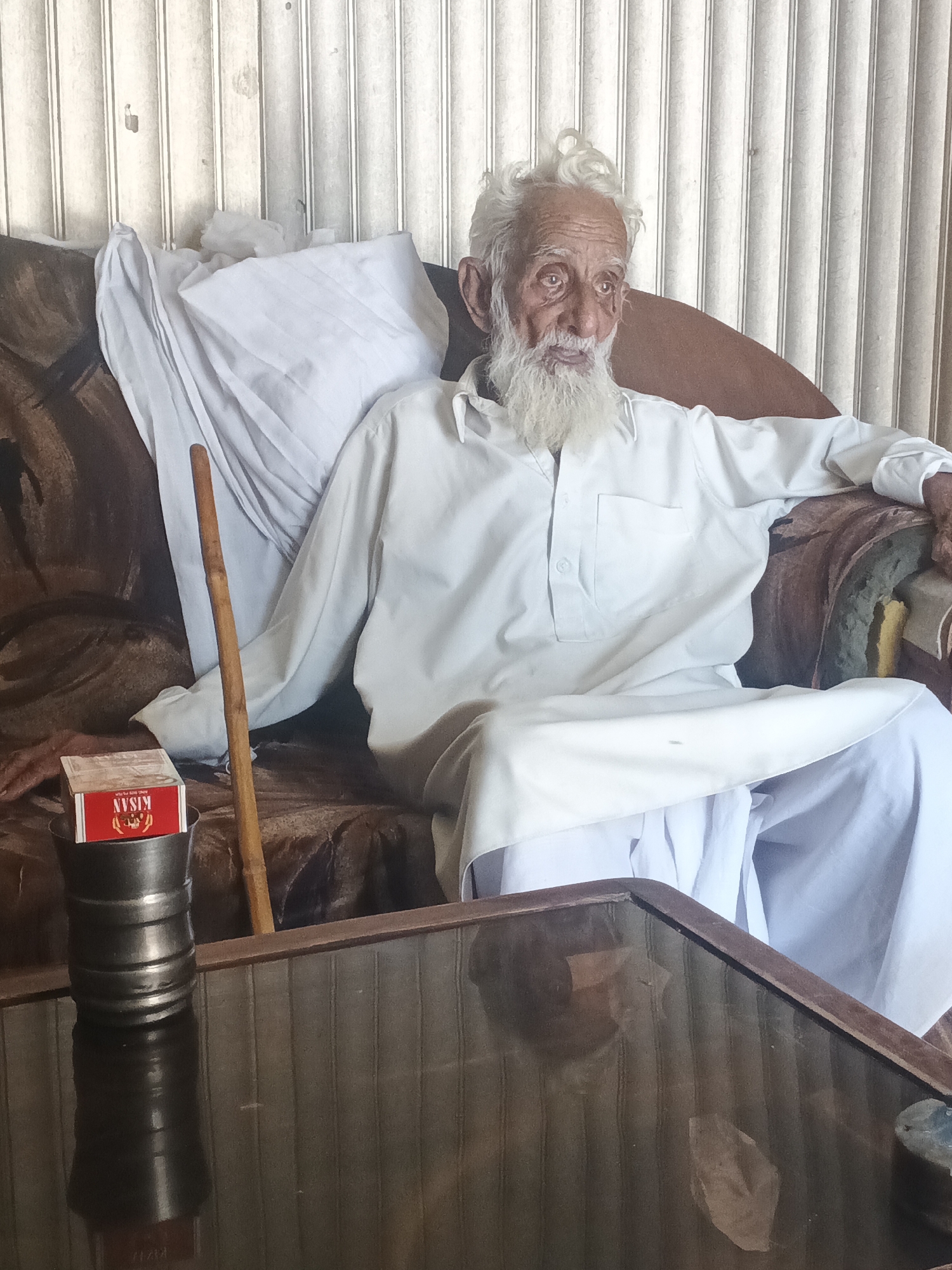 An elderly man with white hair and a long white beard sits on a brown sofa with a white cushion and pillow. He wears a white kurta shalwar and rests his right arm on the back of the sofa. A wooden walking stick leans against the sofa beside him. On a dark, reflective table in front of him is a metal container holding a red box of Kisan brand matches. The background shows vertically striped walls. The photo was taken in Samundri, Pakistan.