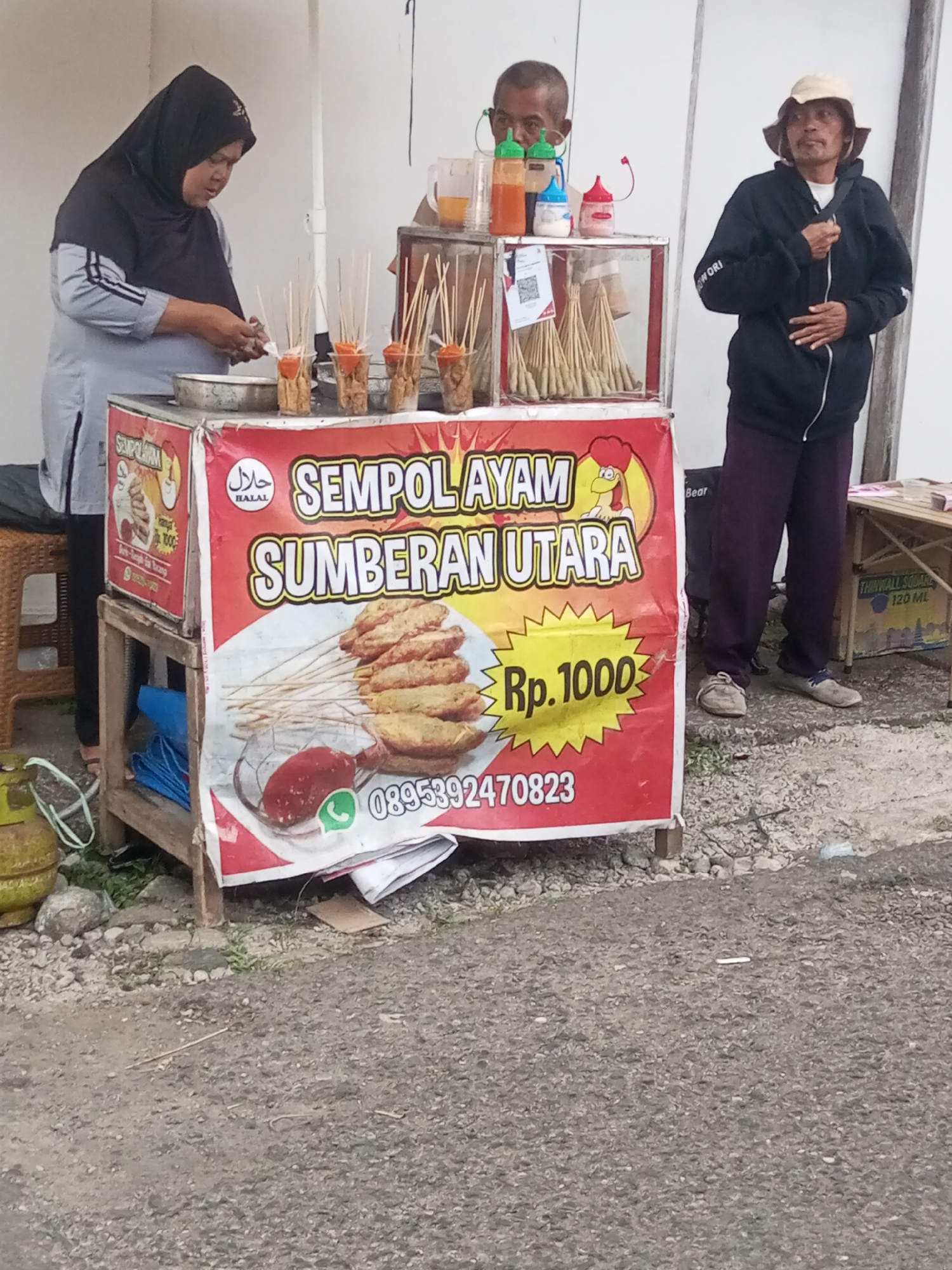 In Pandansari, Indonesia, a street food stall vendor, a woman wearing a black hijab and a grey shirt, prepares food. Beside her, a man with glasses, wearing a black jacket and a hat, stands behind the stall. Another man, also wearing a black jacket and a hat, stands to the right of the stall. The stall displays various food items on skewers, alongside bottles of condiments. A red banner advertises "Sempul Ayam Sumberan Utara" with a price of "Rp. 1000" and a phone number. The stall is situated on a paved road with a background of a white wall.